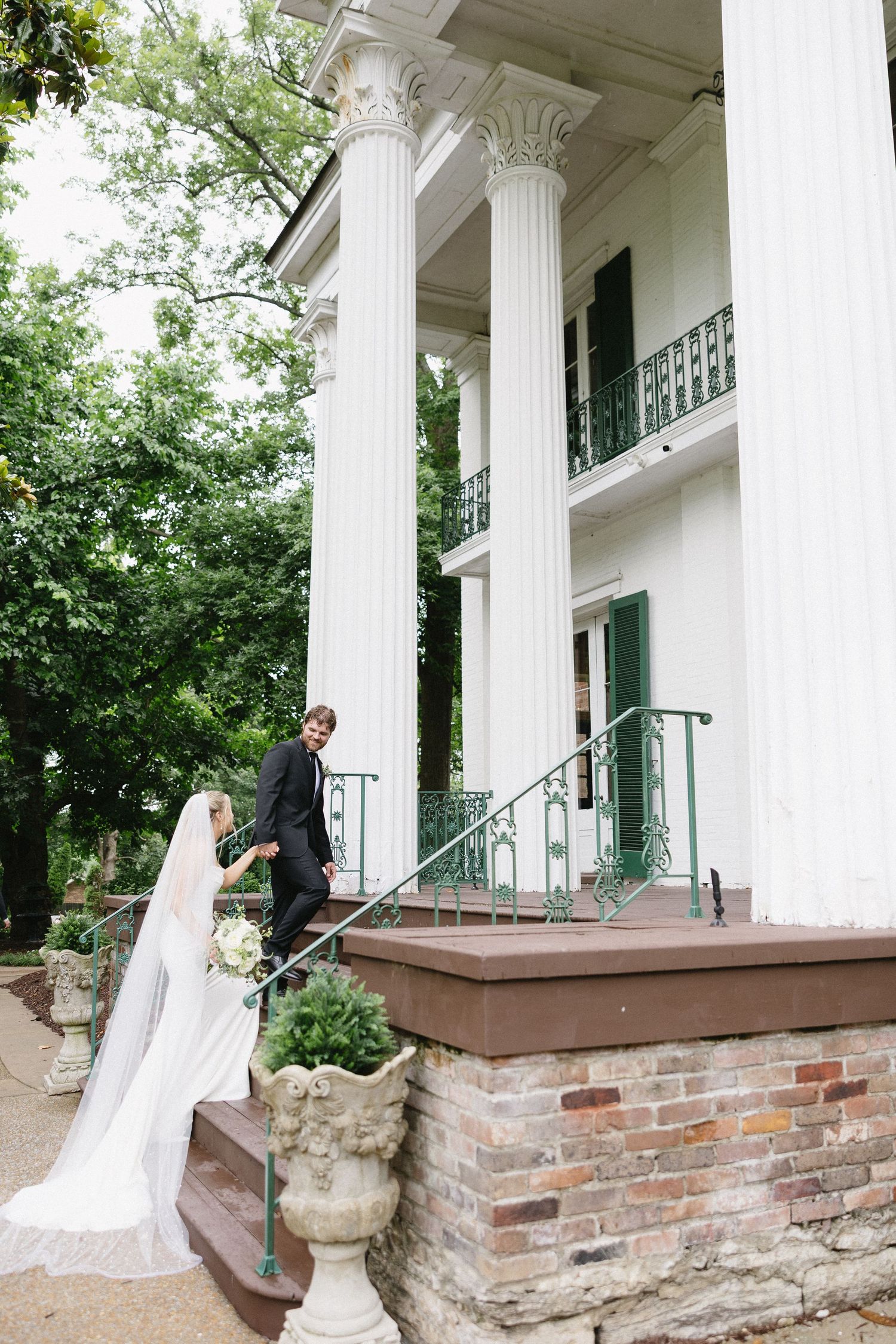 Bride and groom hold hands on steps of elegant Southern antebellum mansion with white columns on their wedding day.