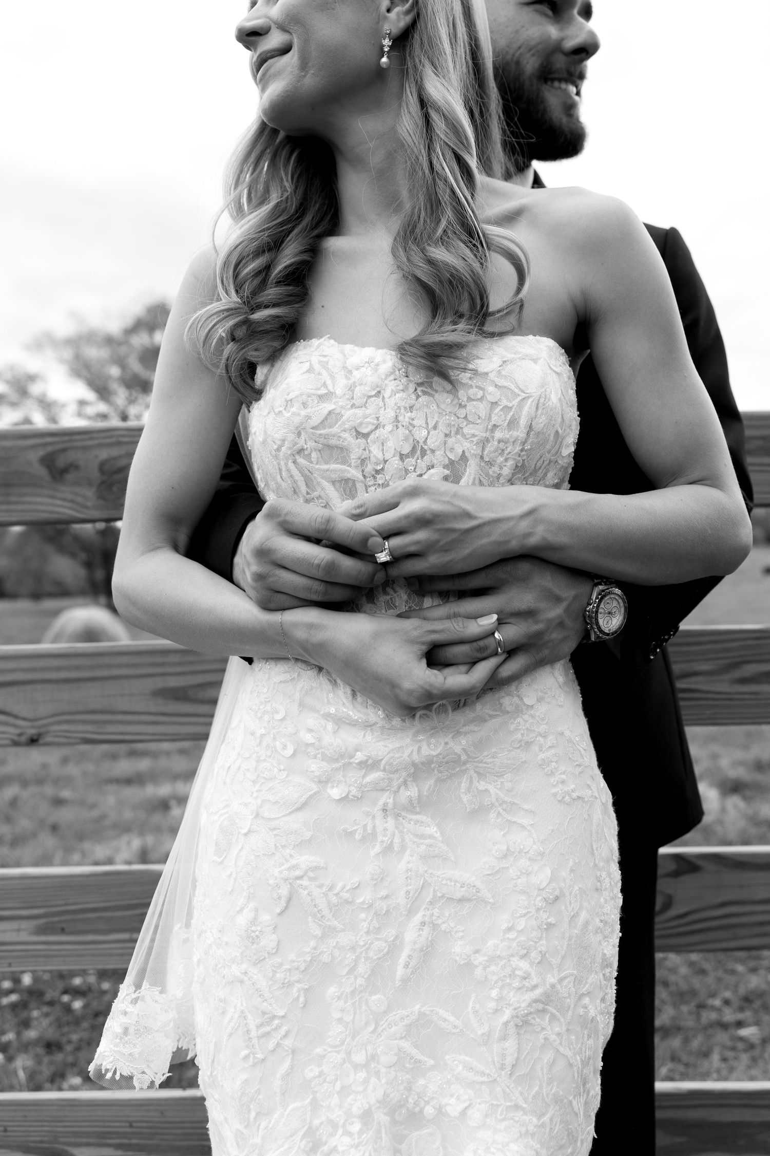Black and white photo of a bride in lace gown embraced by groom in suit, both smiling, near a wooden fence.