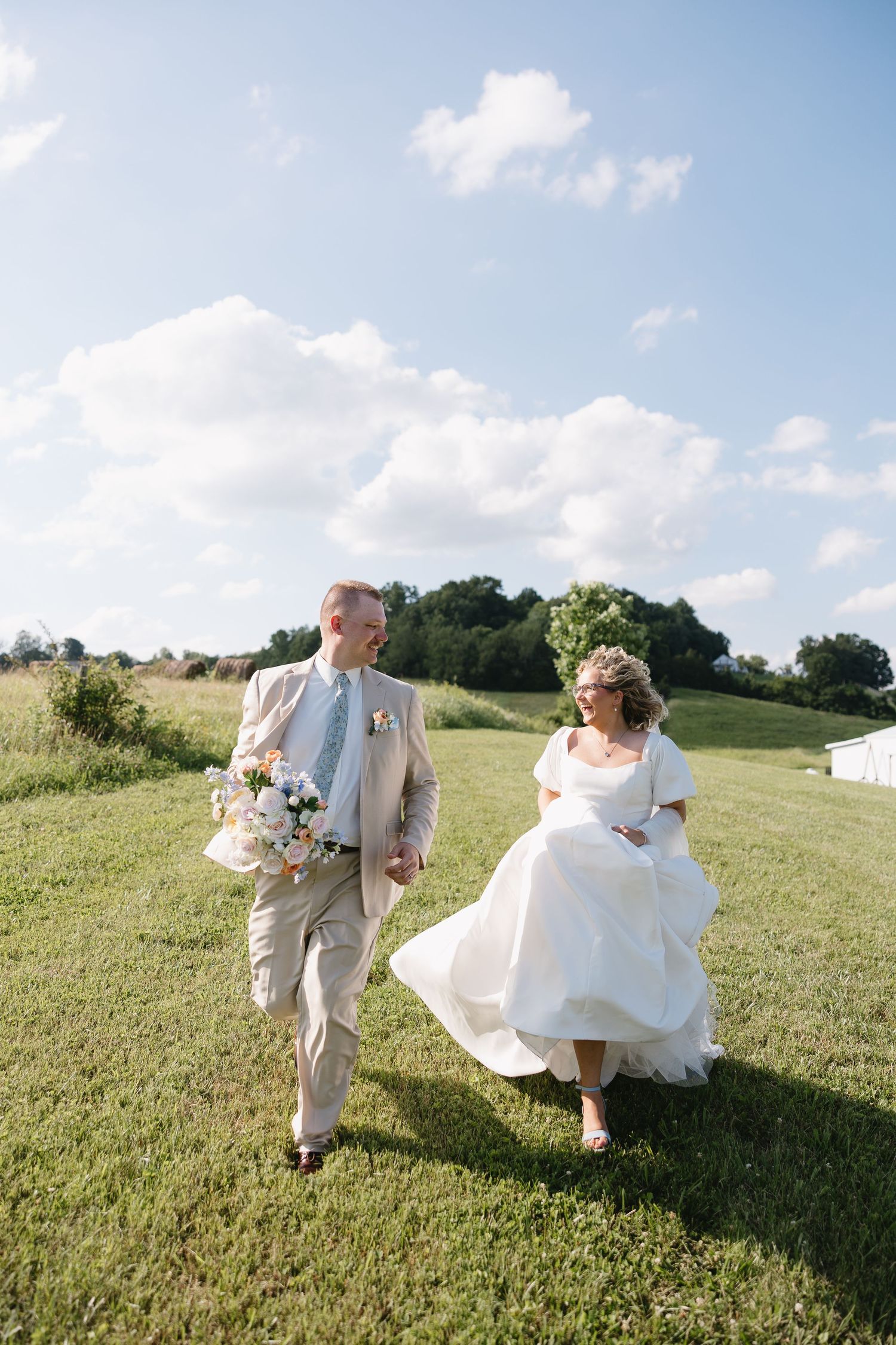 Bride and groom walk hand-in-hand across green meadow, bride in white gown holding bouquet on sunny wedding day.