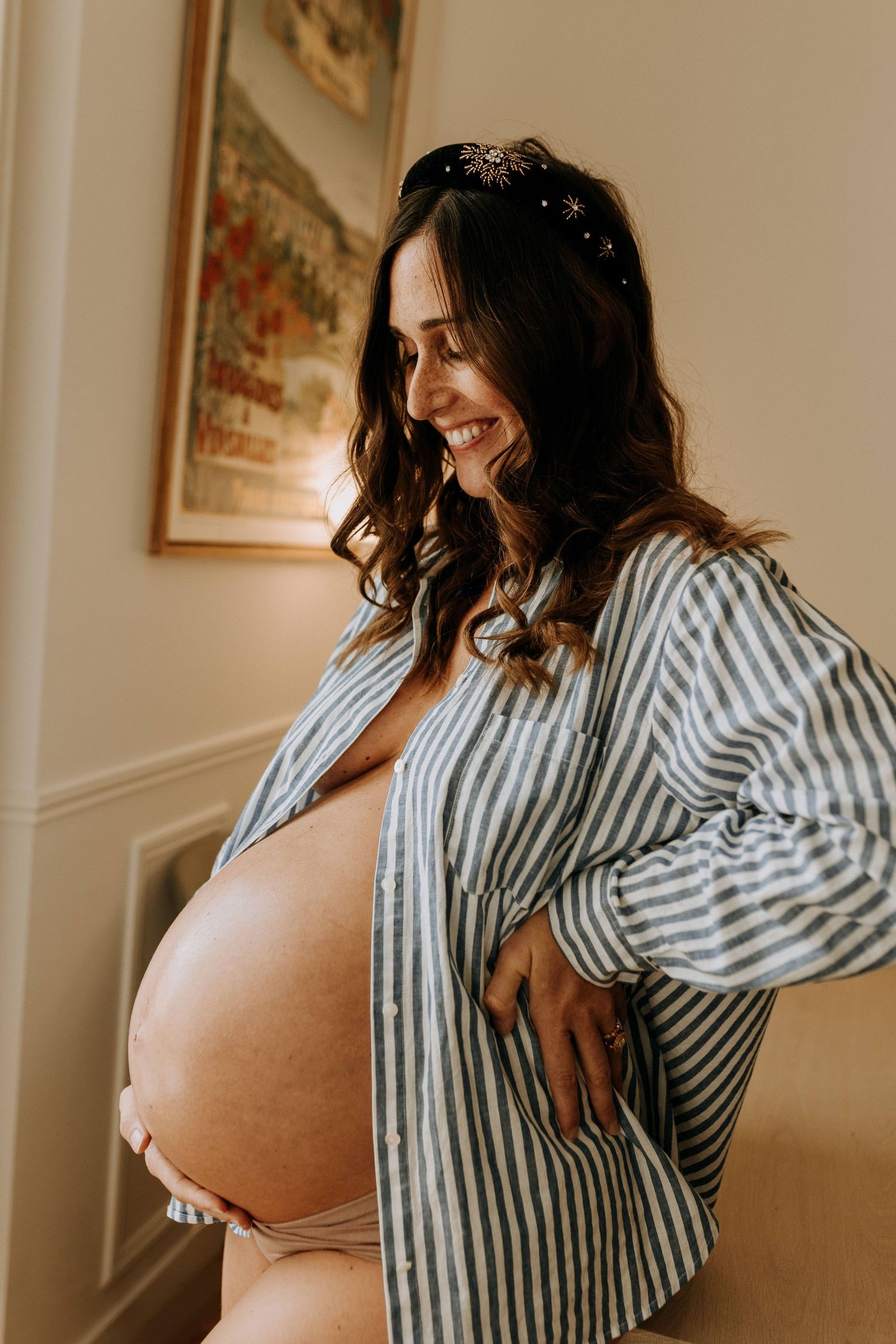 Séance photo grossesse à domicile : future maman souriante, ventre rond mis en valeur, lumière naturelle et moment doux capturé sur le vif.