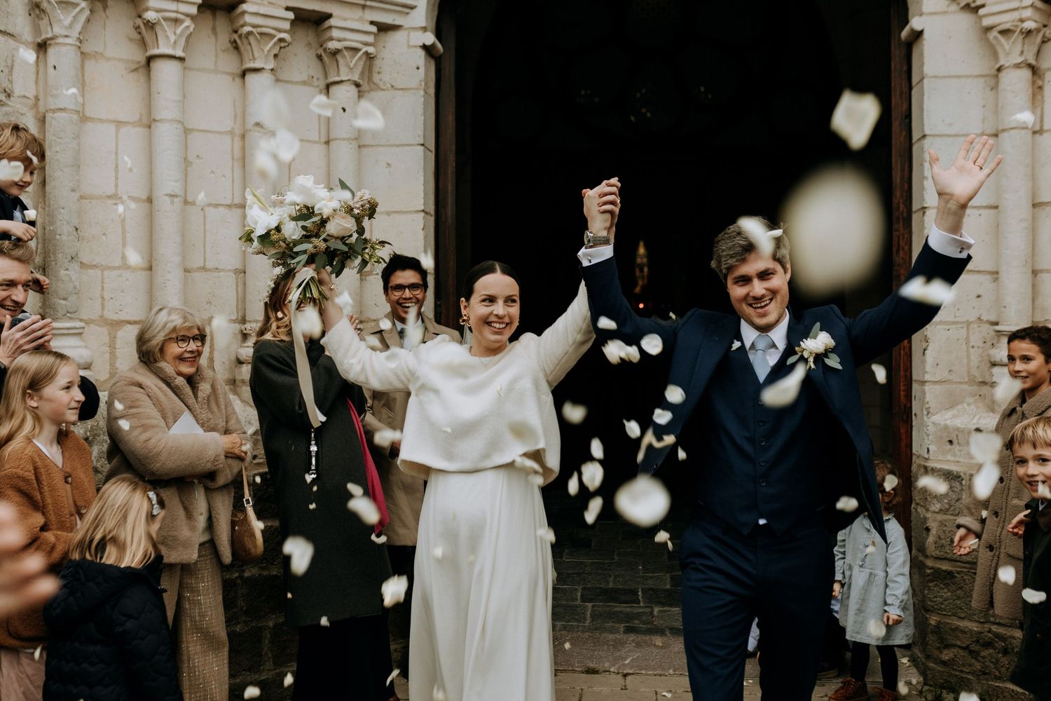 Couple de mariés sortant de l’église sous une pluie de pétales, sourires et émotions partagées entourés de leurs proches.