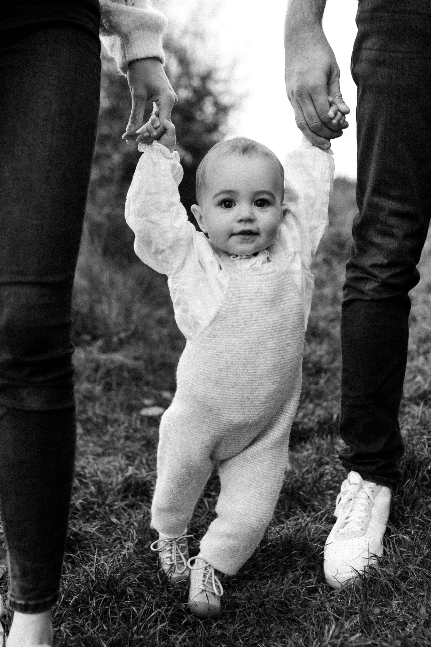 Bébé tenu par les mains de ses parents lors d’une séance photo famille en noir et blanc, moment de tendresse authentique