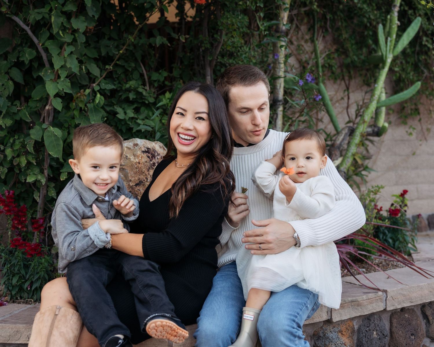 A family sits together outdoors on wooden steps surrounded by lush greenery and flowers in a casual photo session.