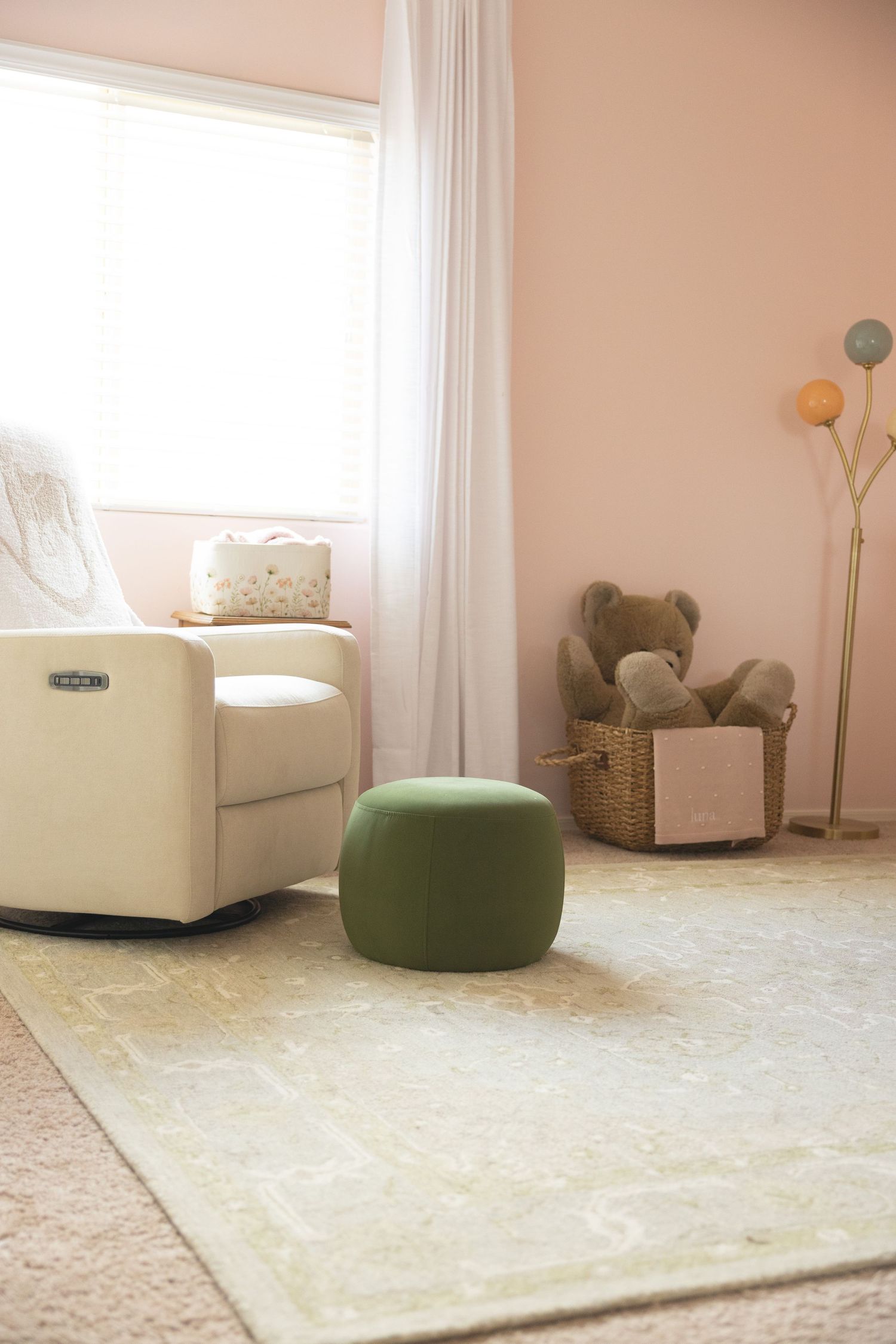 Soft pink nursery with cream glider, green ottoman, vintage rug, and teddy bear in wicker basket.