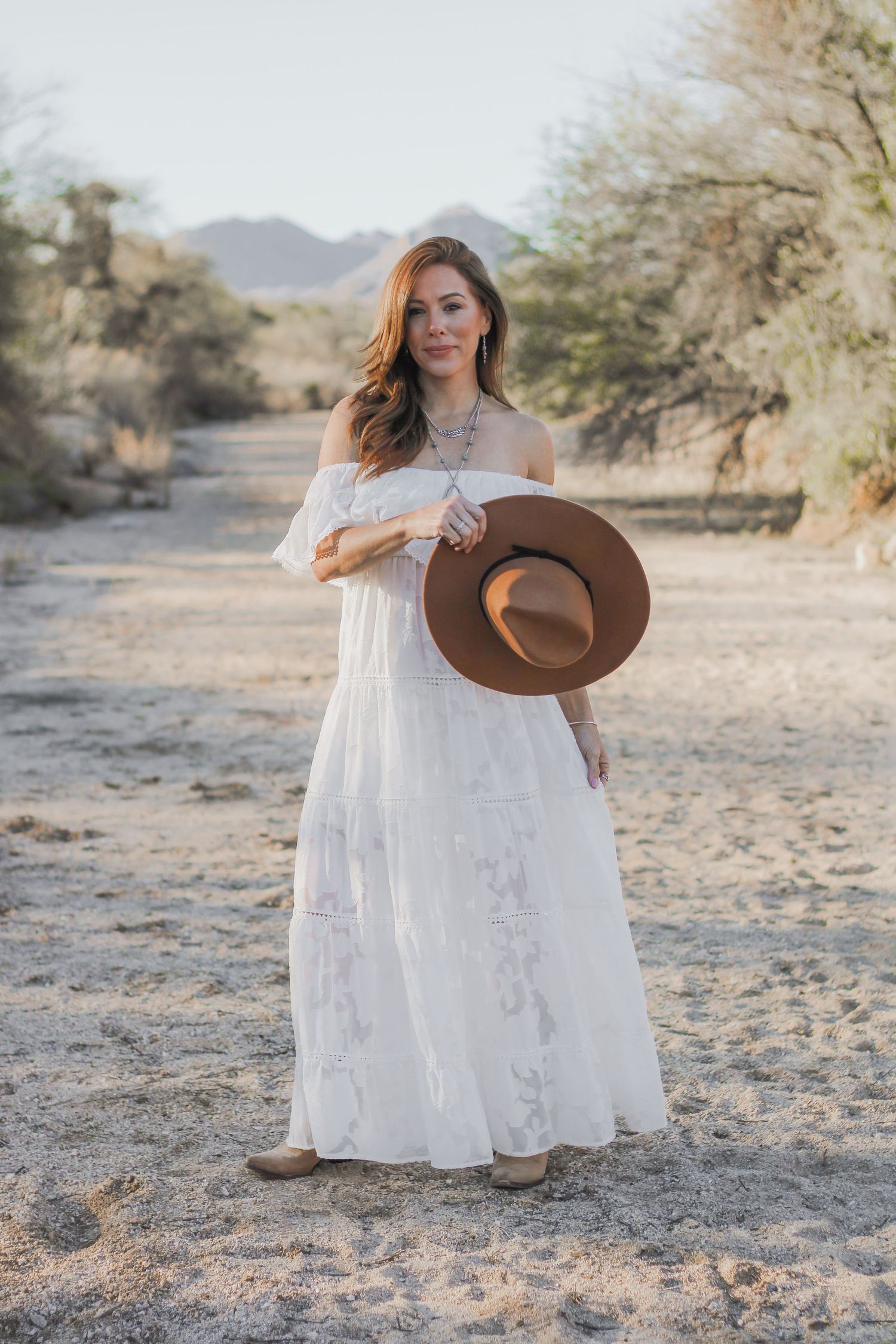 Woman in white lace dress holding brown hat in desert landscape during golden hour.