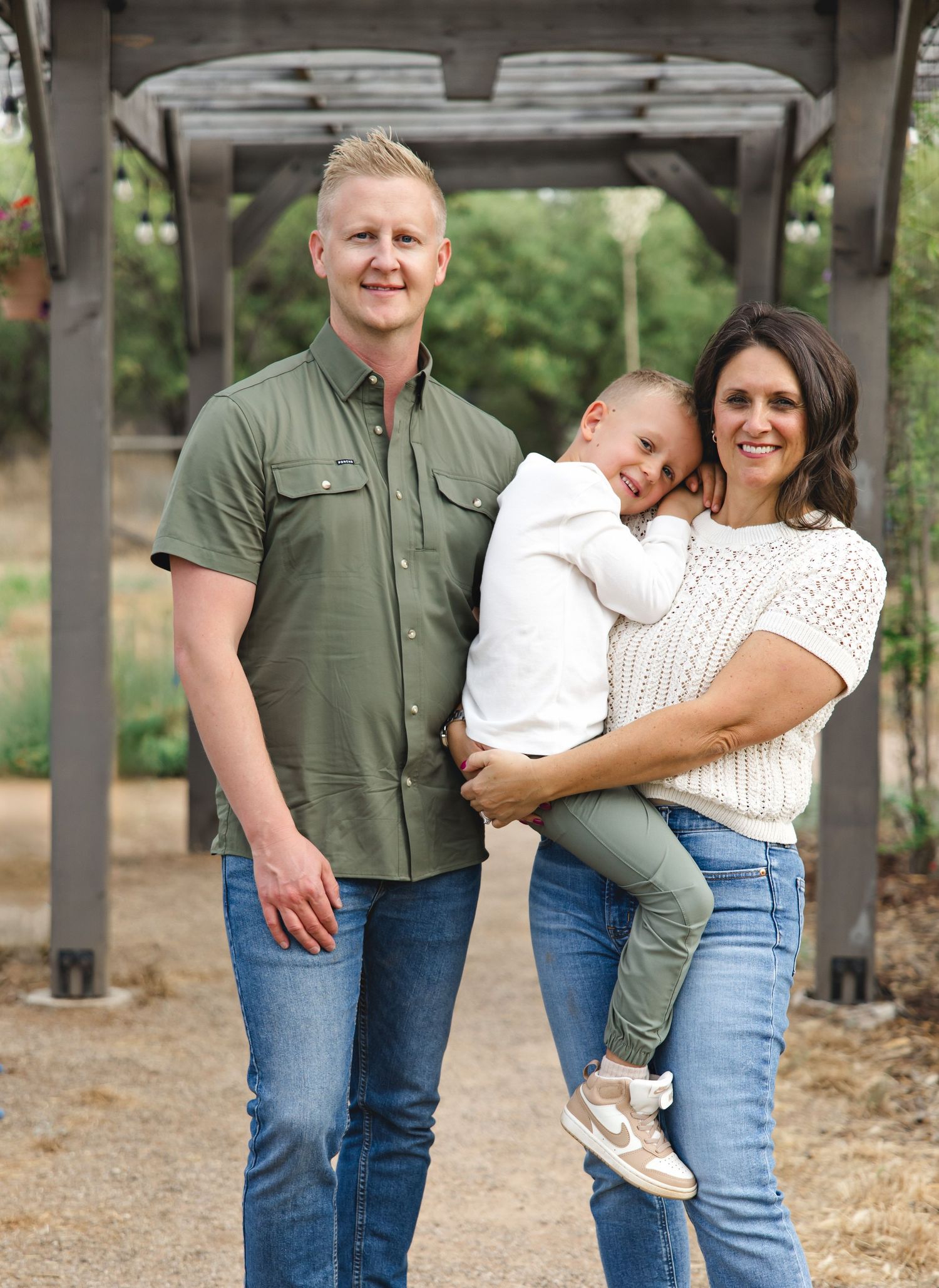 Smiling family of three stands outdoors under a wooden pergola, with a man in a green shirt, a woman in a white knit top, and a young child.