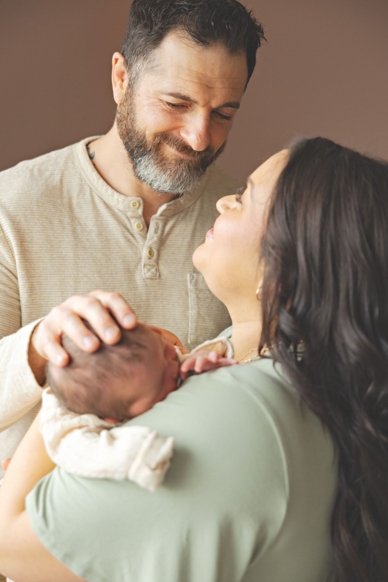 Smiling bearded father and brunette mother gazing at each other while holding their newborn baby.