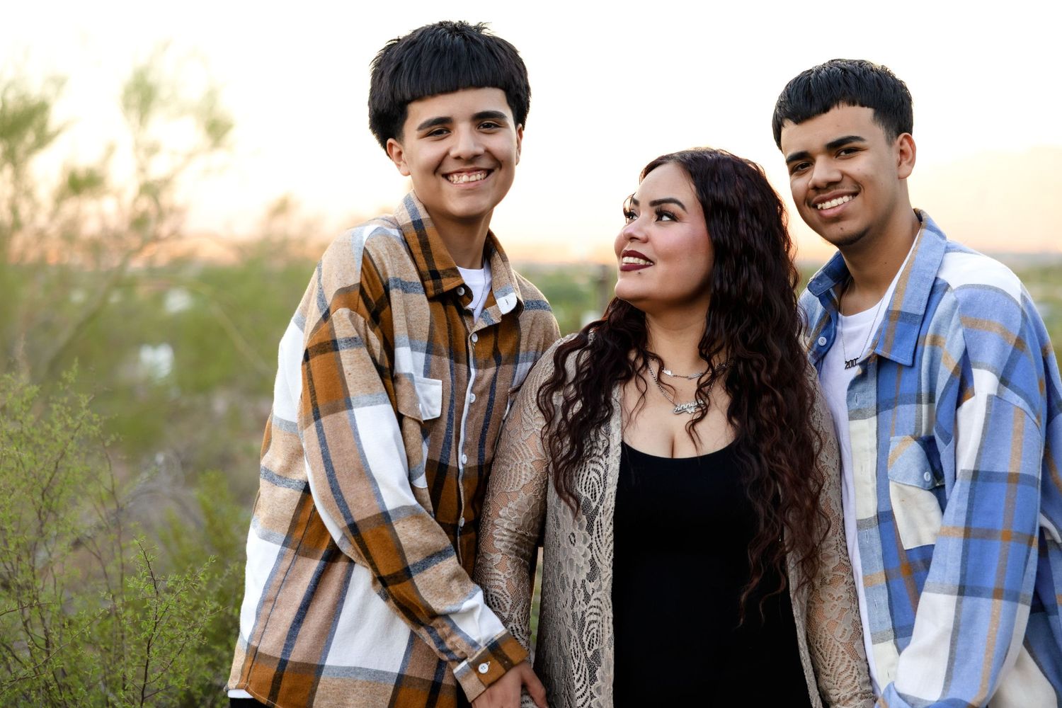 Three young friends in plaid shirts and casual wear smile together outdoors during golden hour.