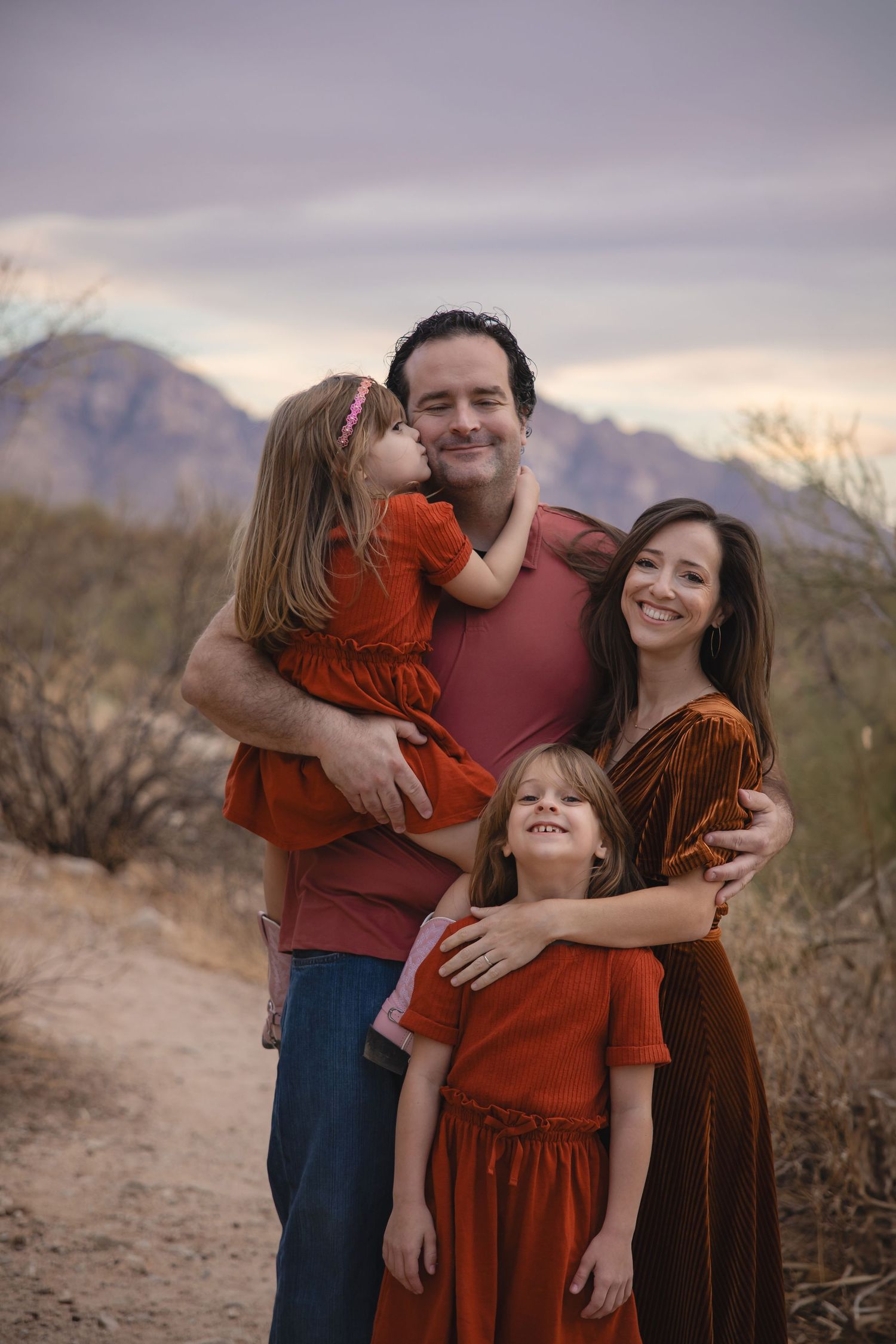 A family wearing coordinating red and burgundy outfits embraces while posing in a desert landscape at sunset.