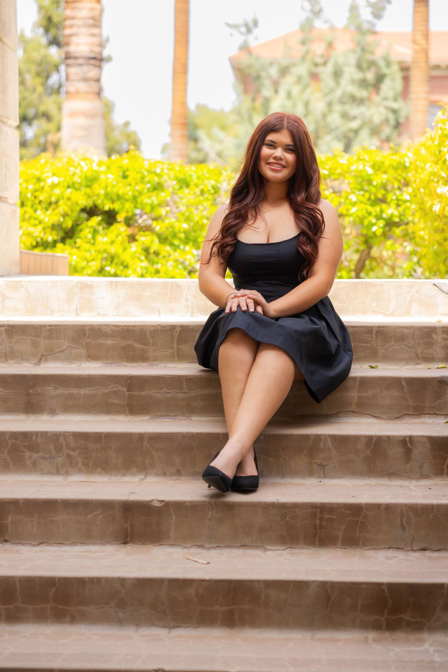 A person in a black dress sits on outdoor stone steps with yellow flowers and palm trees in the background.
