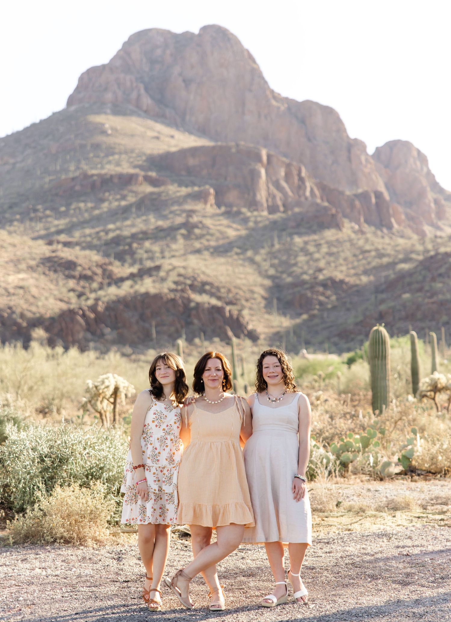 Three women in summer dresses pose together on a desert trail with saguaro cacti and a rugged mountain behind them.