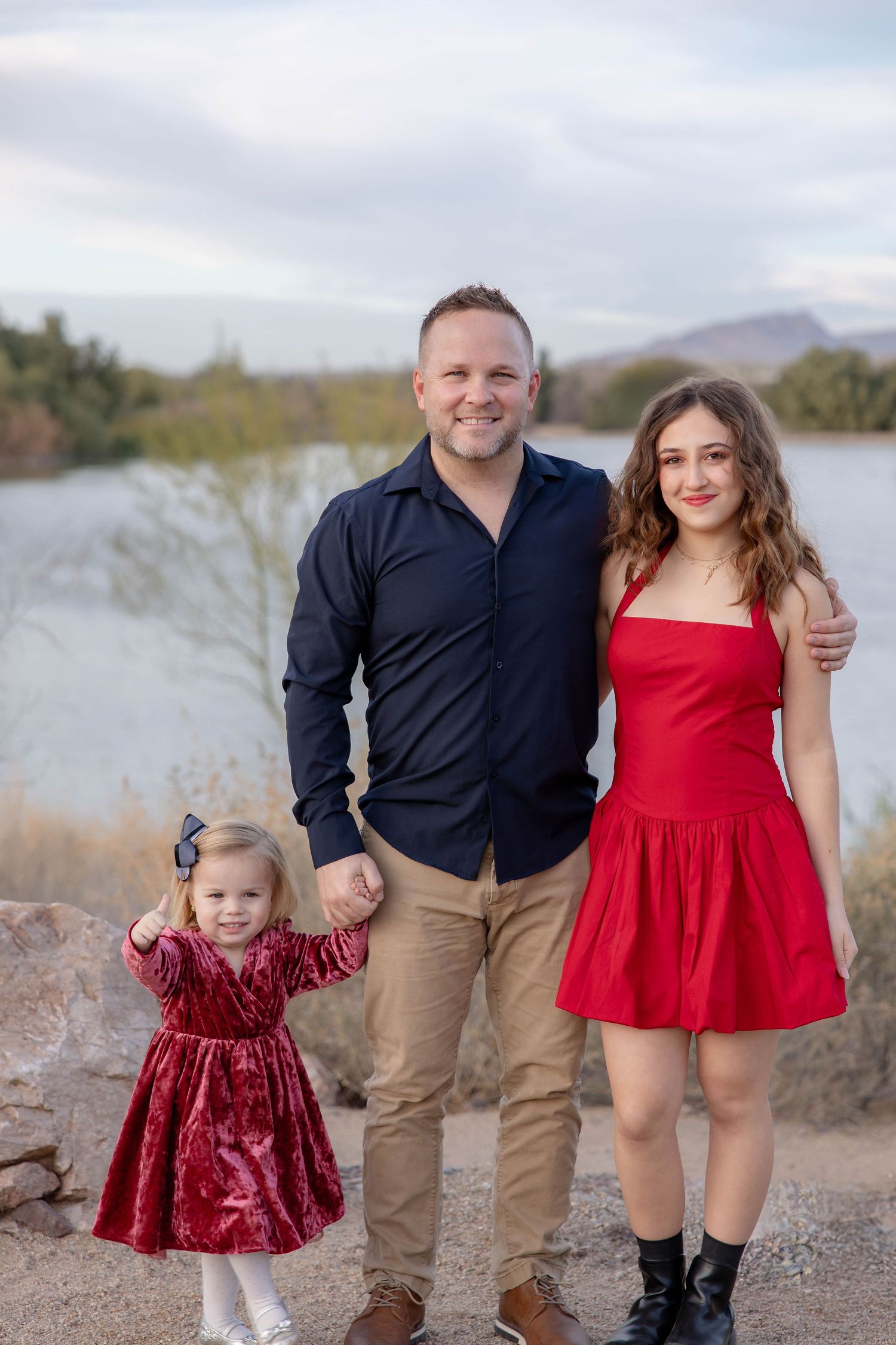 A family in coordinating red and navy outfits poses together on a desert path near a lake during golden hour.