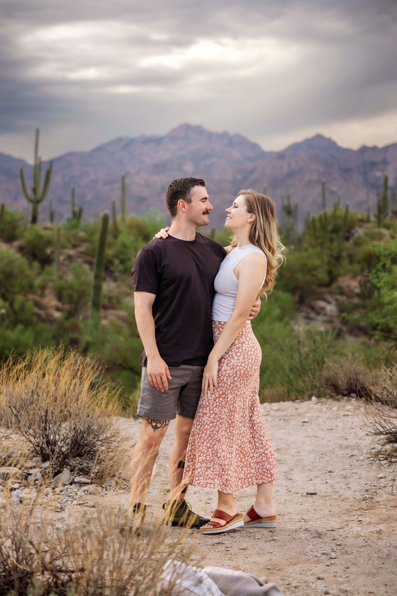 A couple shares a romantic moment on a hiking trail surrounded by desert landscape and mountains.