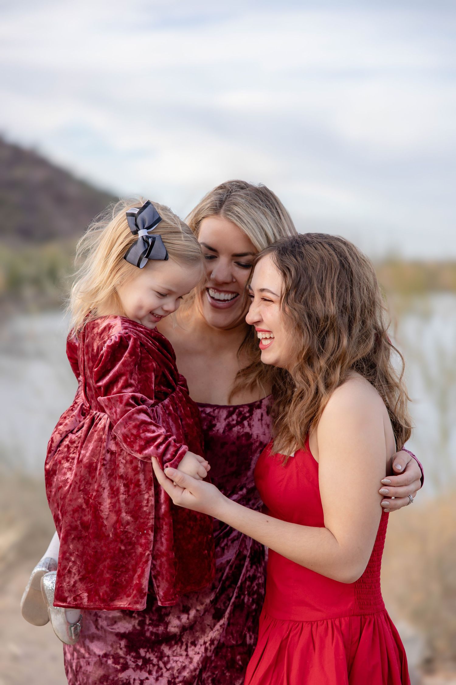 Three people in red dresses share a joyful moment together outdoors against a natural landscape.