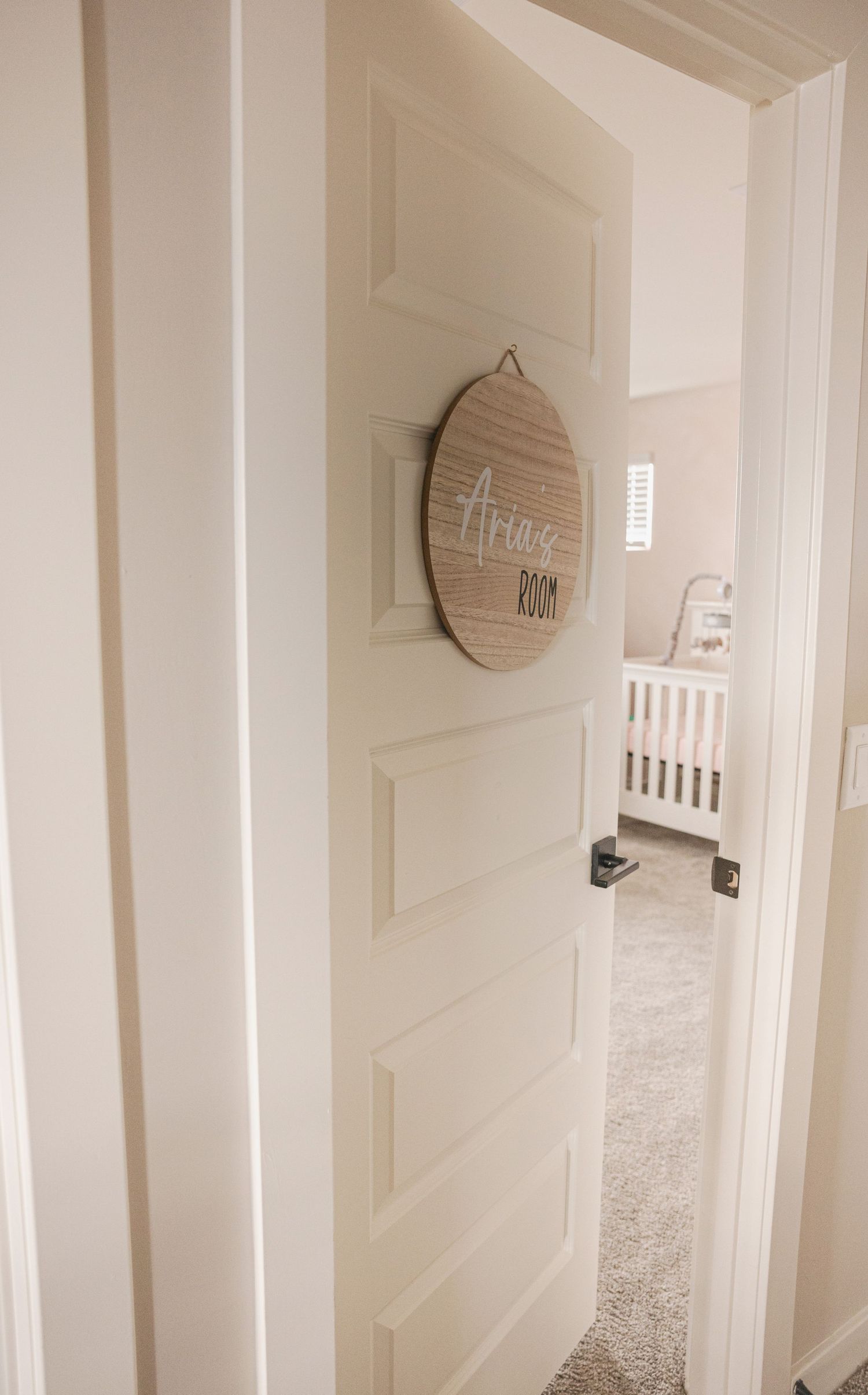 A nursery hallway with a wooden round sign reading Aria's Room on a white door, with a white crib visible inside.