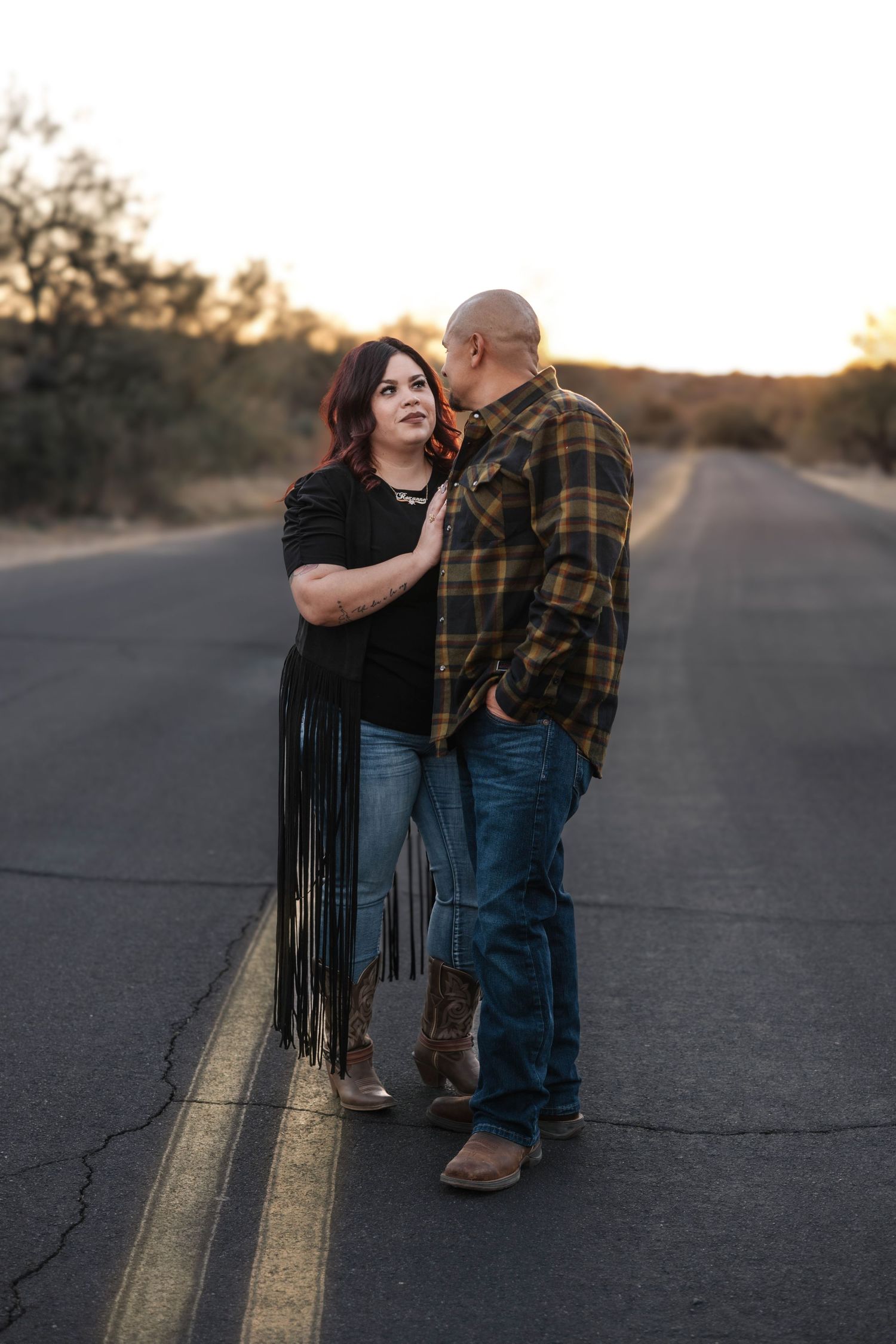 A couple shares an intimate moment on a rural road at sunset as shadows stretch across the pavement.