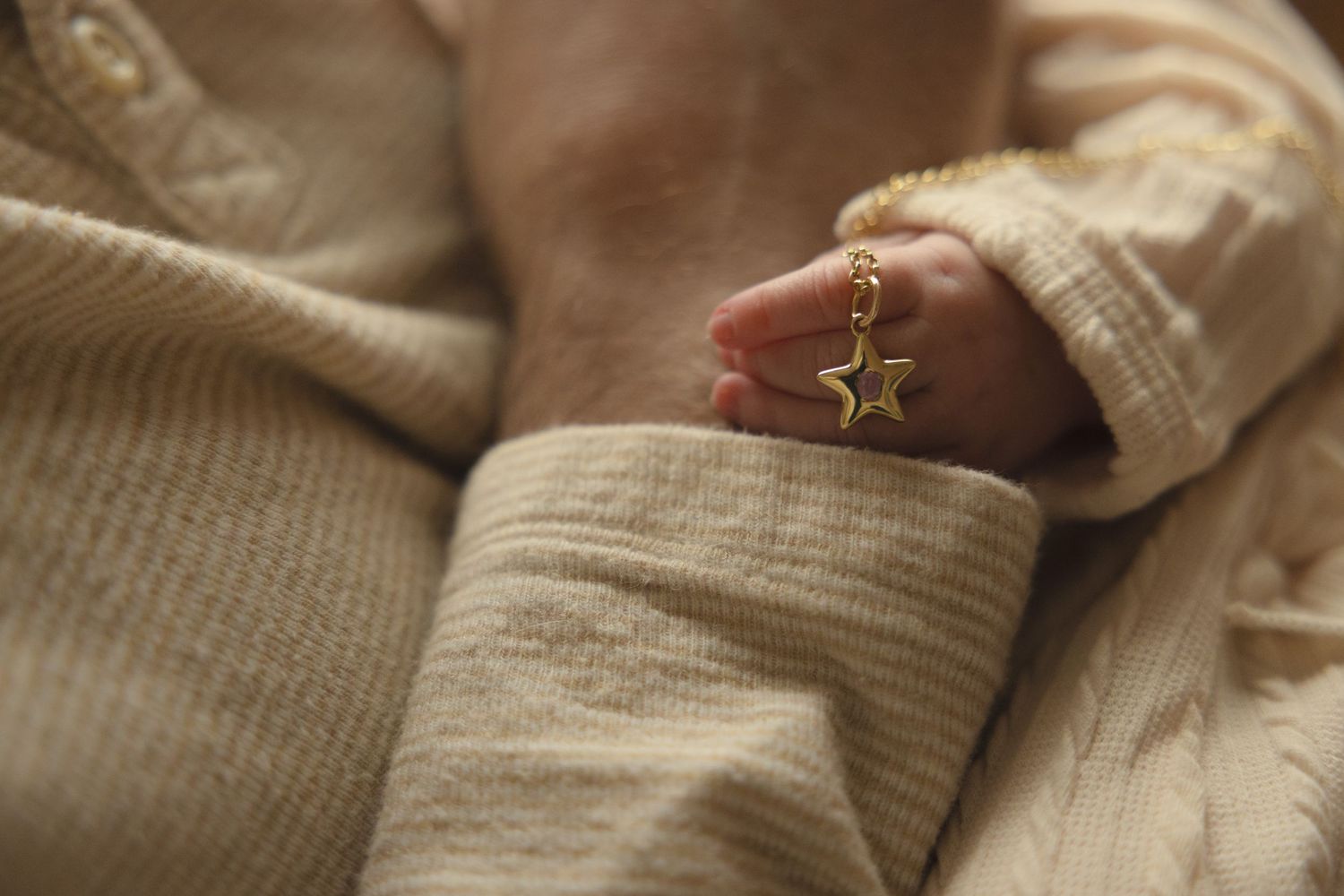 Newborn baby hand clutching a gold star charm necklace in a cream knit outfit.