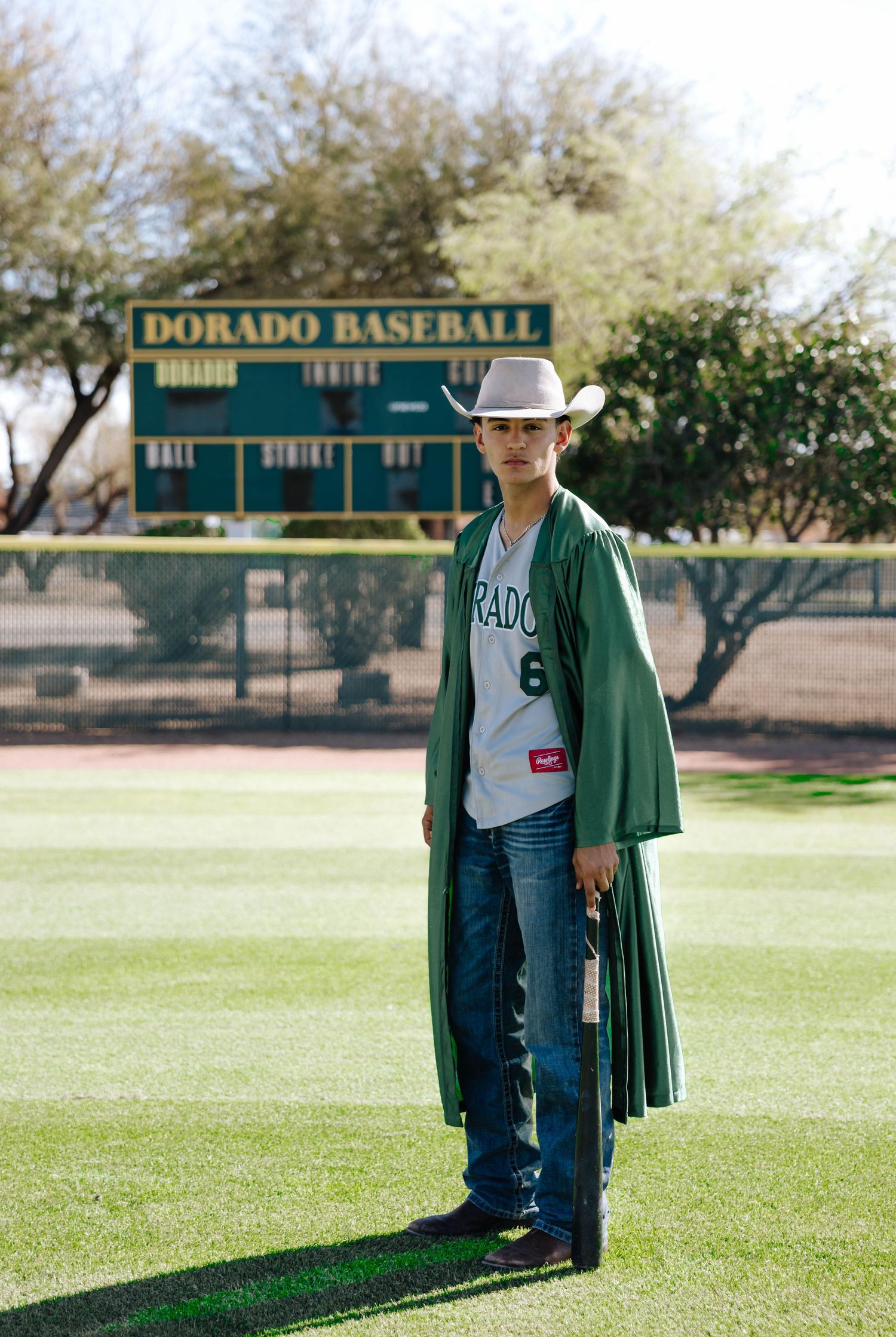 Baseball player in green jacket stands on sunny field with Dorado Baseball sign visible in background.