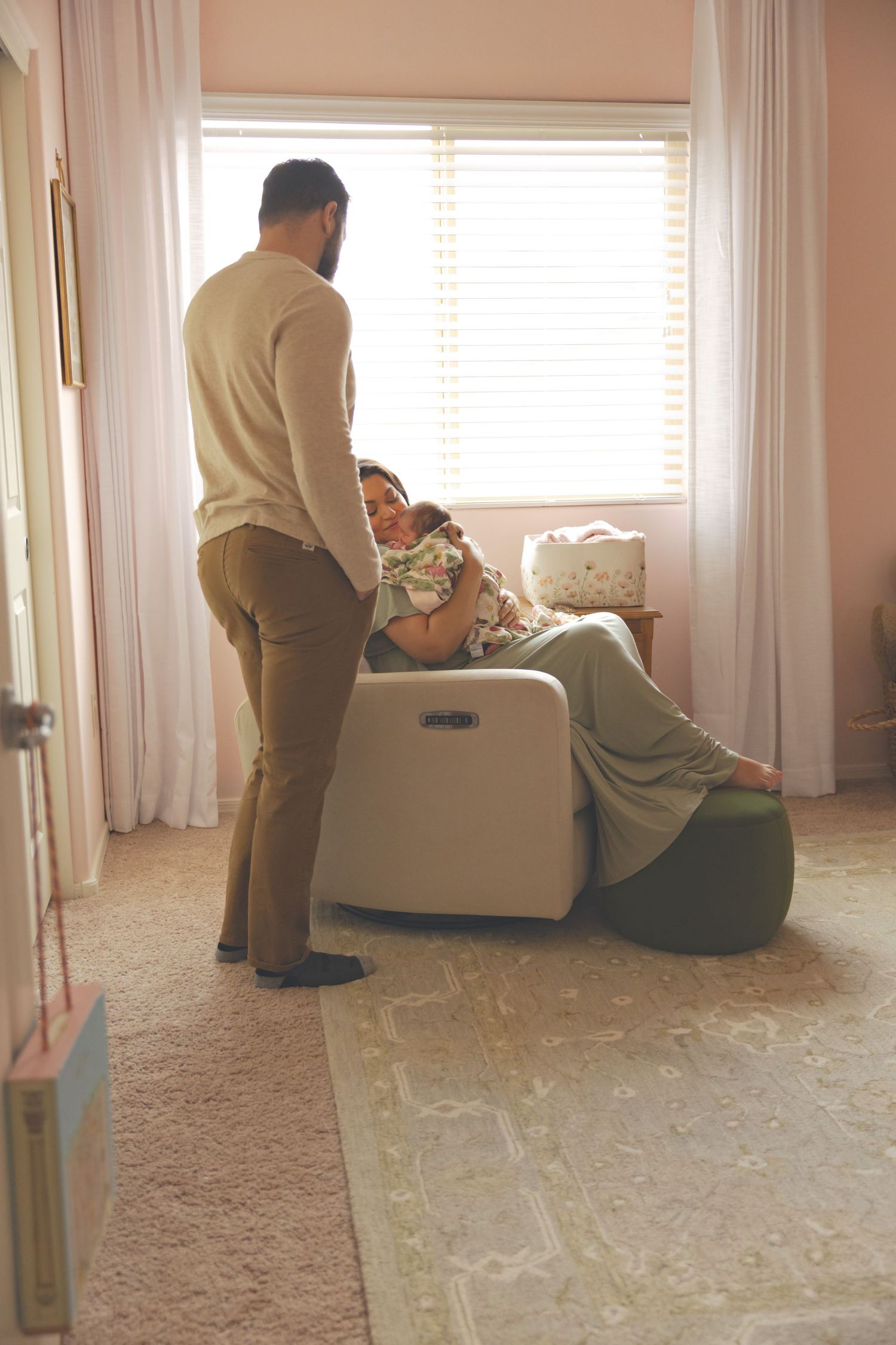 A couple in a softly lit nursery, with a woman reclining in a white chair holding a newborn as her partner stands beside her.