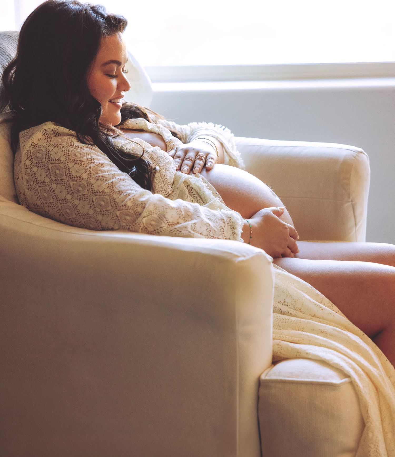 Pregnant woman in lace robe relaxing in a cream armchair, cradling her baby bump in soft natural light.