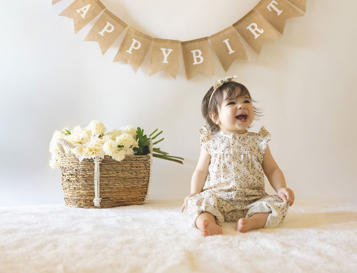 Happy baby girl in a floral romper sits laughing on a white fluffy rug beside a basket of white flowers under a burlap birthday banner.