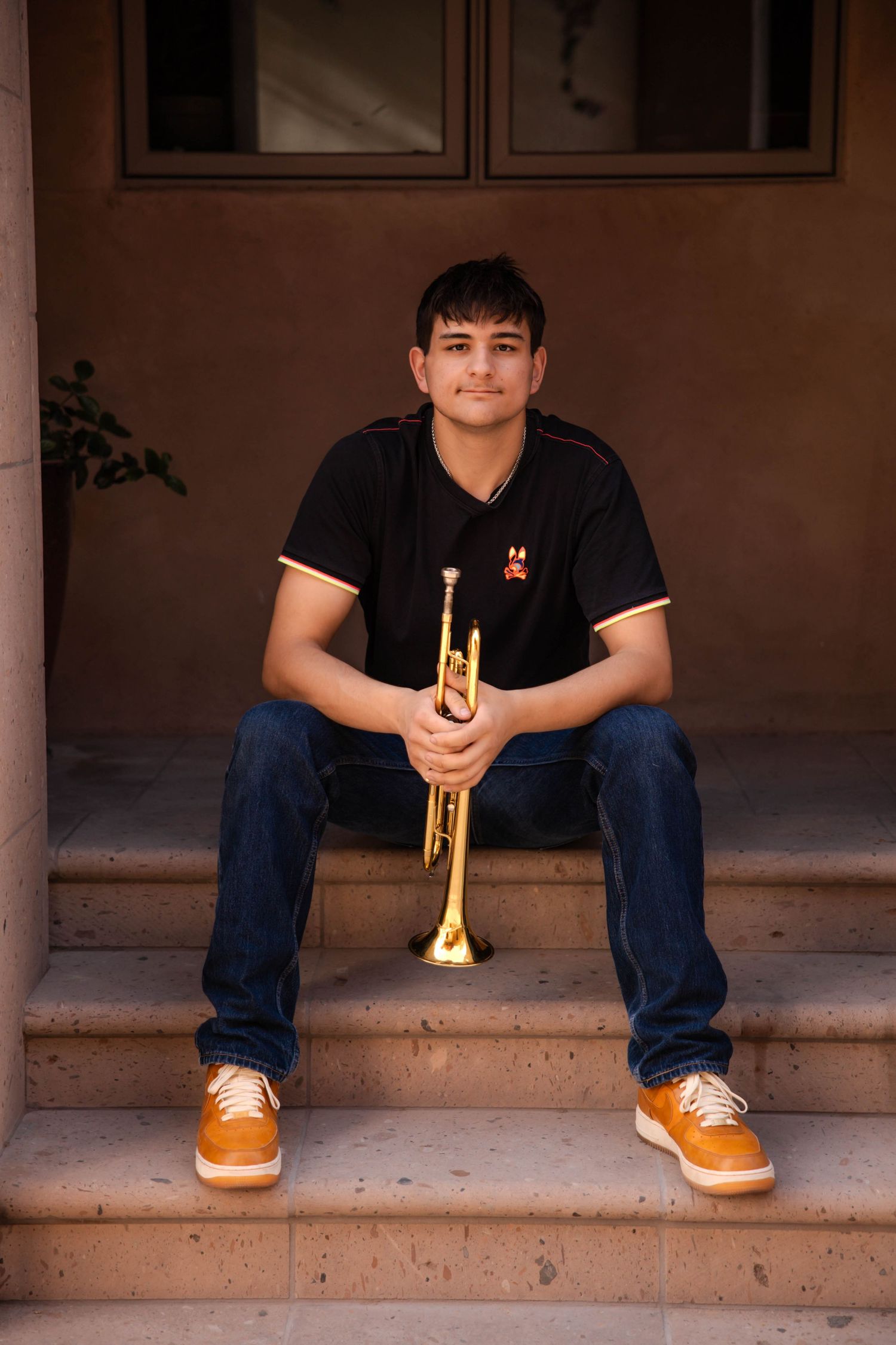 Student sits on porch steps holding a brass trumpet while wearing casual attire and tan shoes.