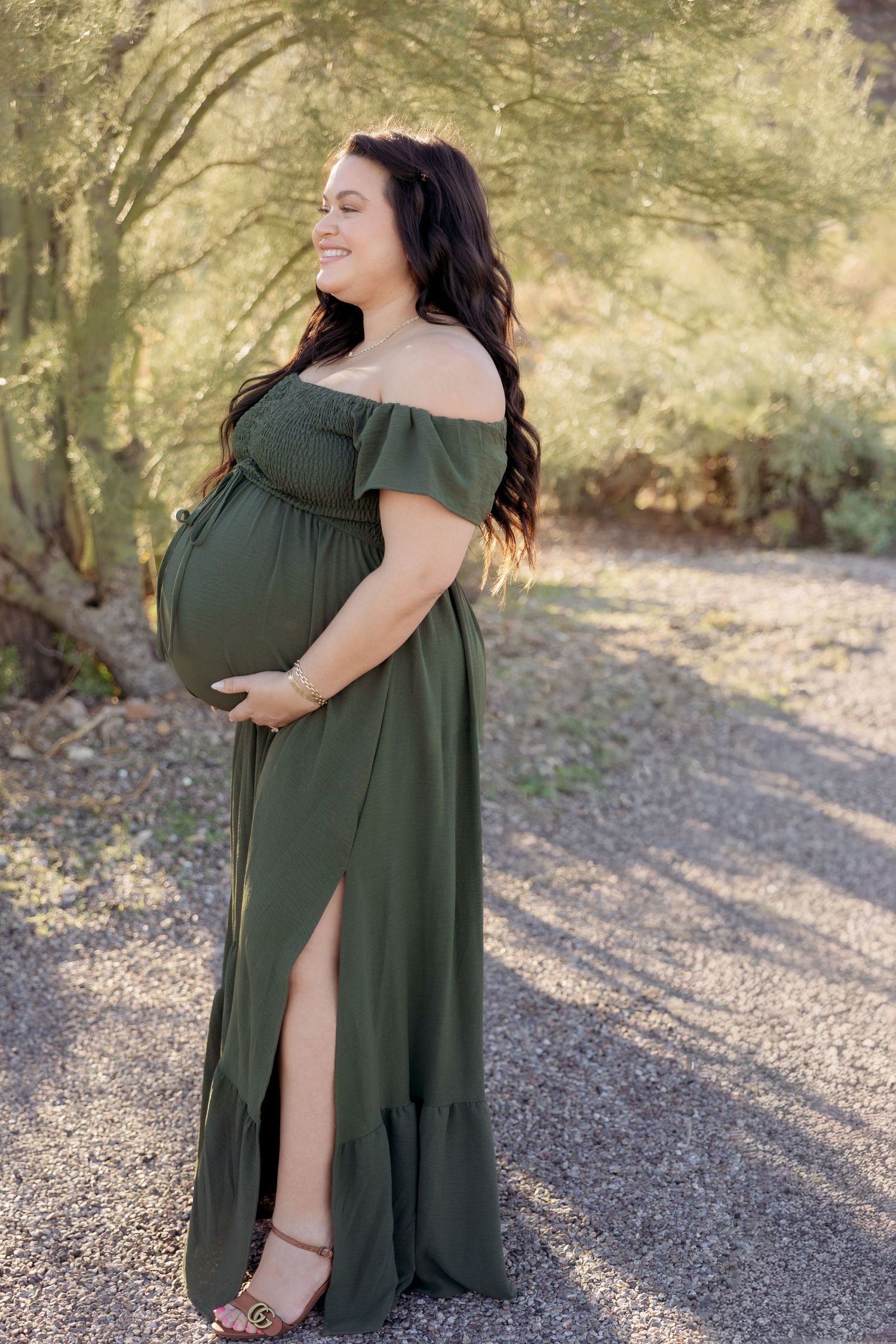 Smiling pregnant woman in olive green off-shoulder maxi dress holding baby bump outdoors on gravel path.