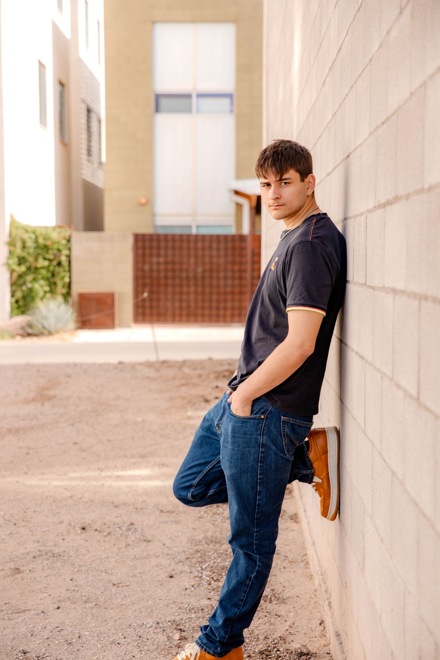 Person in black t-shirt and jeans leaning against white wall in outdoor hallway setting.