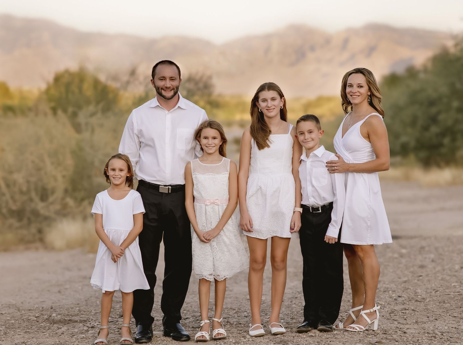 A family of six wearing coordinated white outfits poses together on a desert path with mountains in the background.