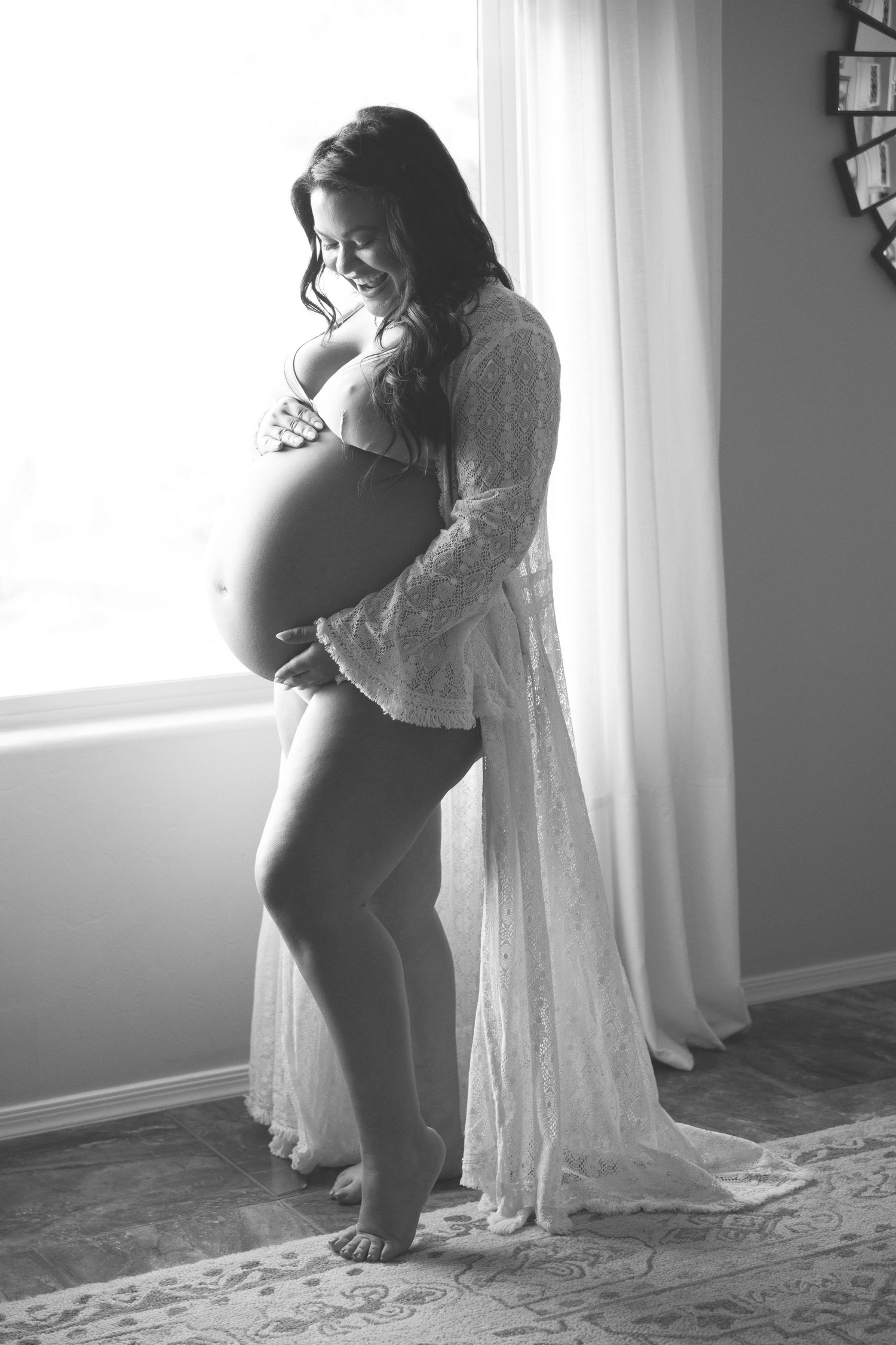 Black and white maternity photoshoot showing silhouette of pregnant woman in flowing dress by window.