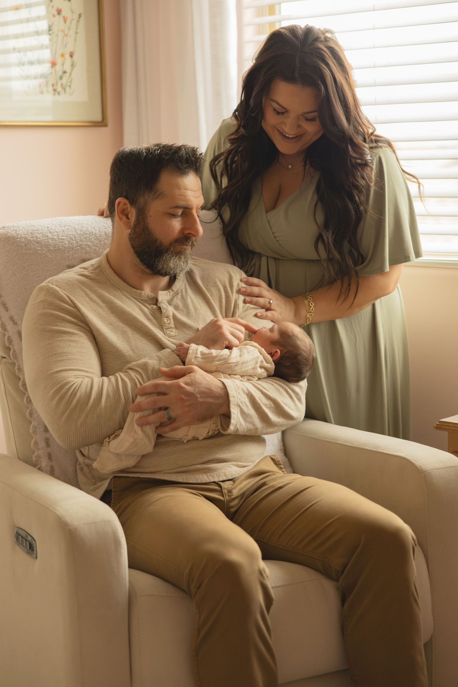 New parents in pink nursery, father seated in cream glider holding newborn while mother stands beside.