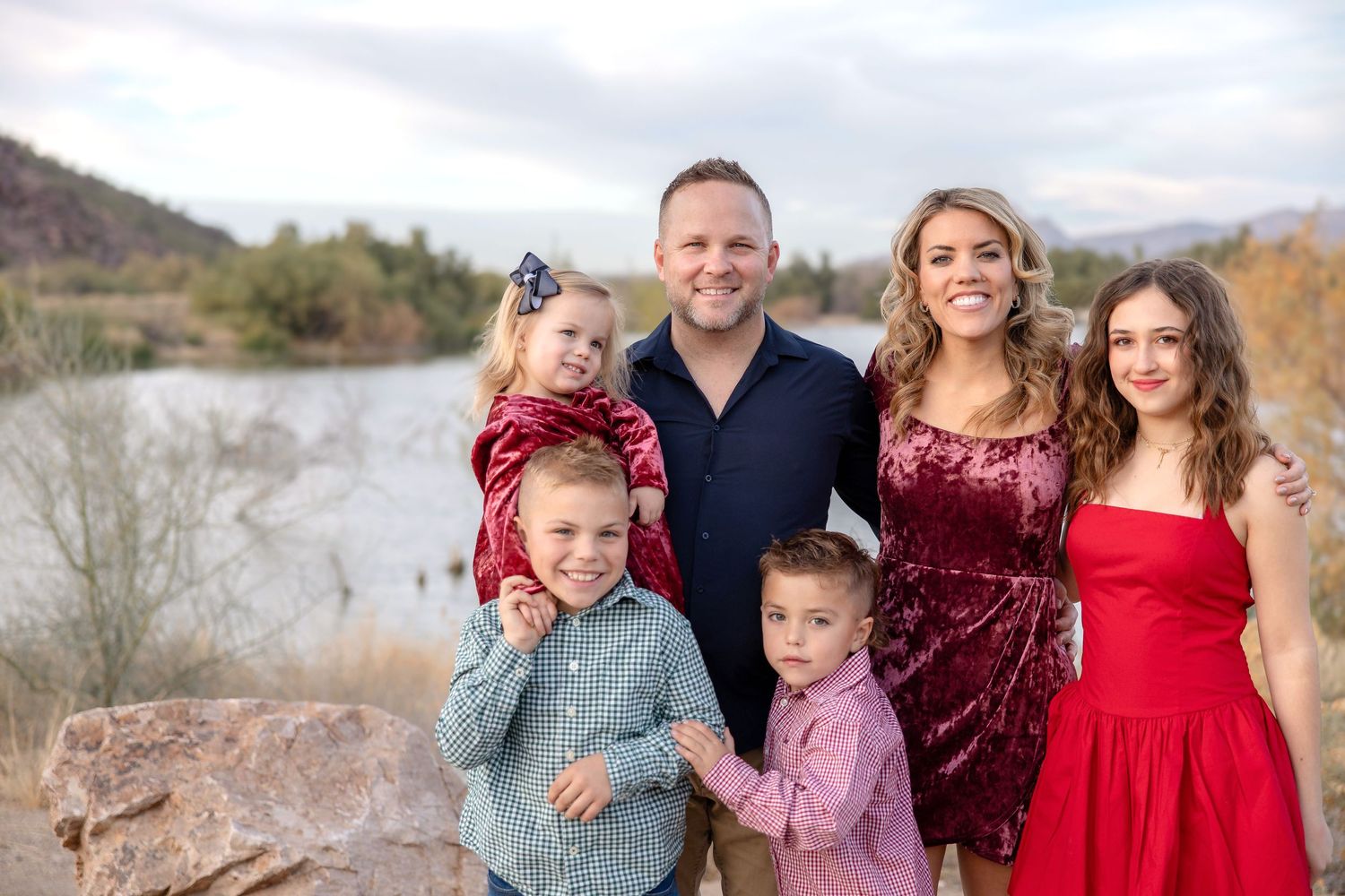 A family wearing coordinated red and burgundy outfits poses together in a scenic outdoor setting.