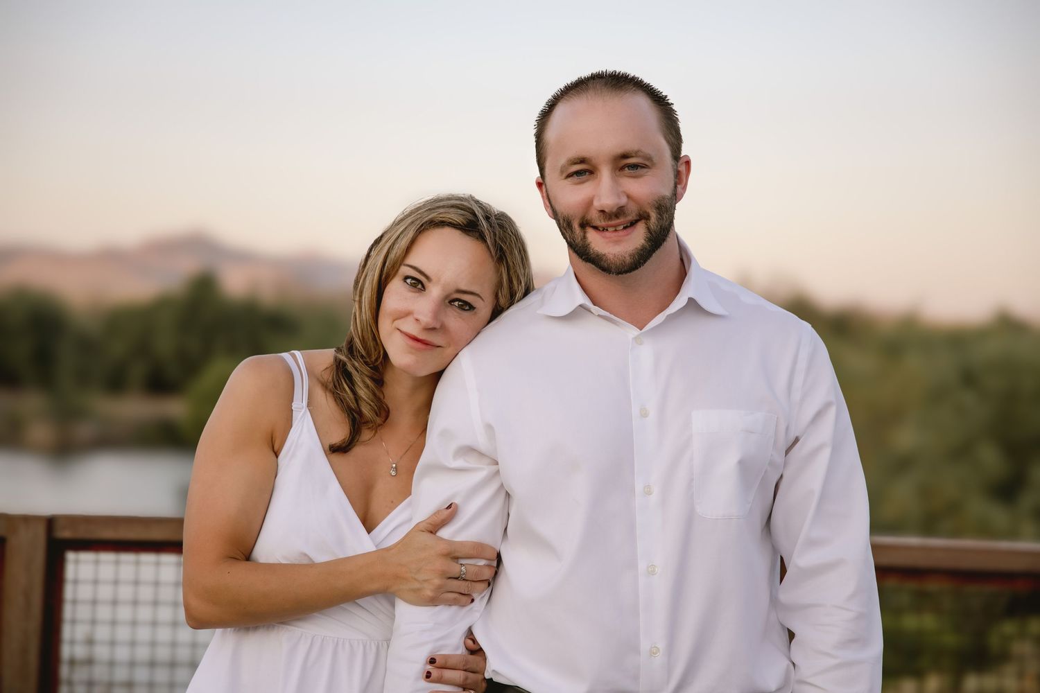 A couple in white outfits embraces outdoors during a sunset photo session in a desert landscape.