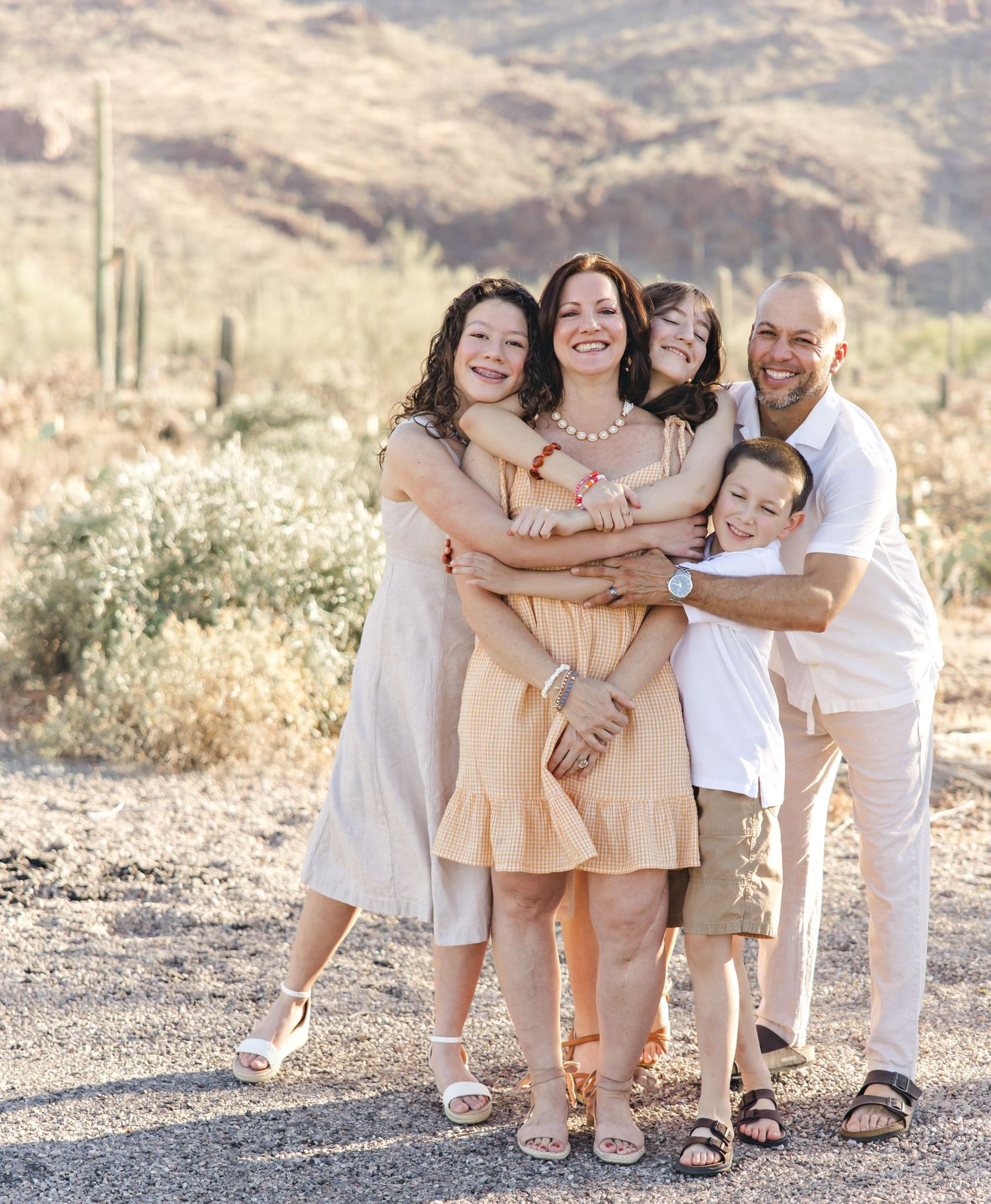 Family of five in neutral outfits embracing and smiling together in a sunny desert landscape with saguaro cacti and hills.