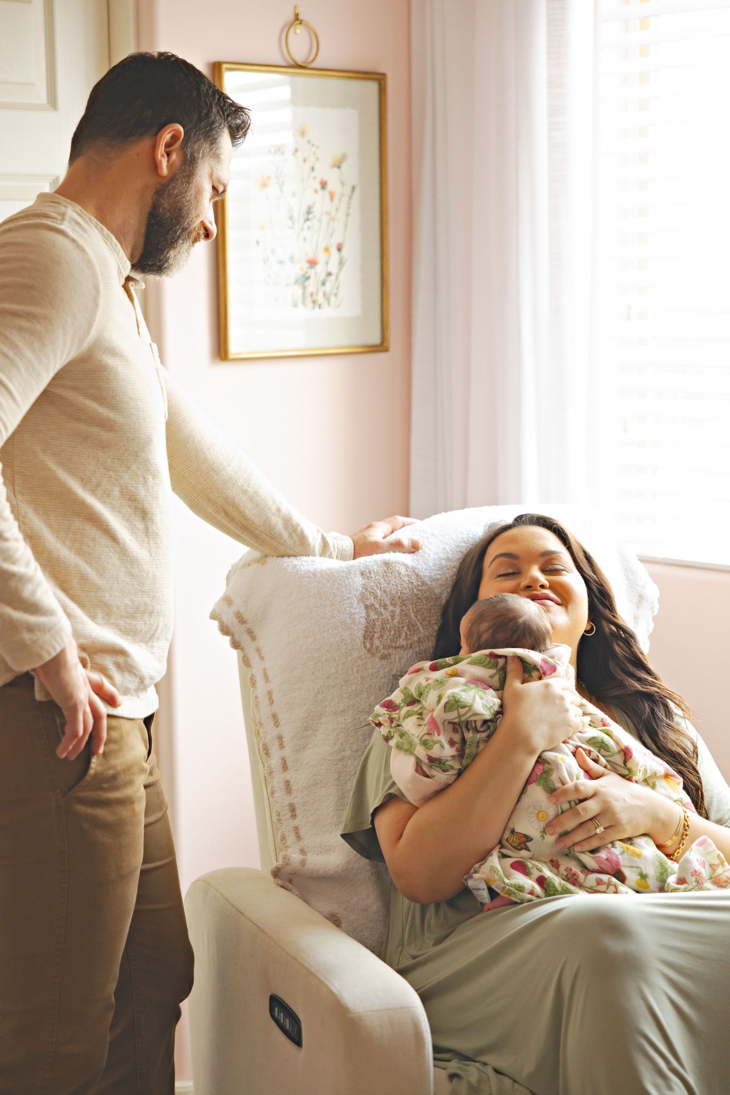 A man stands beside a woman sitting in a nursery chair, smiling as she holds their newborn baby in soft natural light.