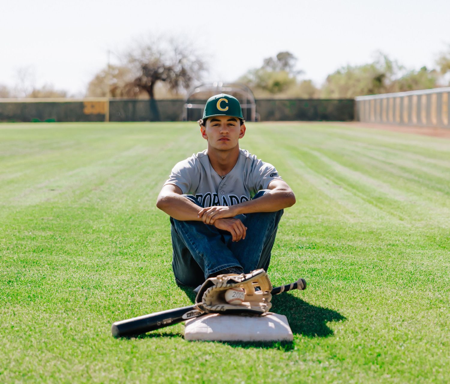 Baseball player in green cap leans on wooden bat in sunny grass field during outdoor practice session.