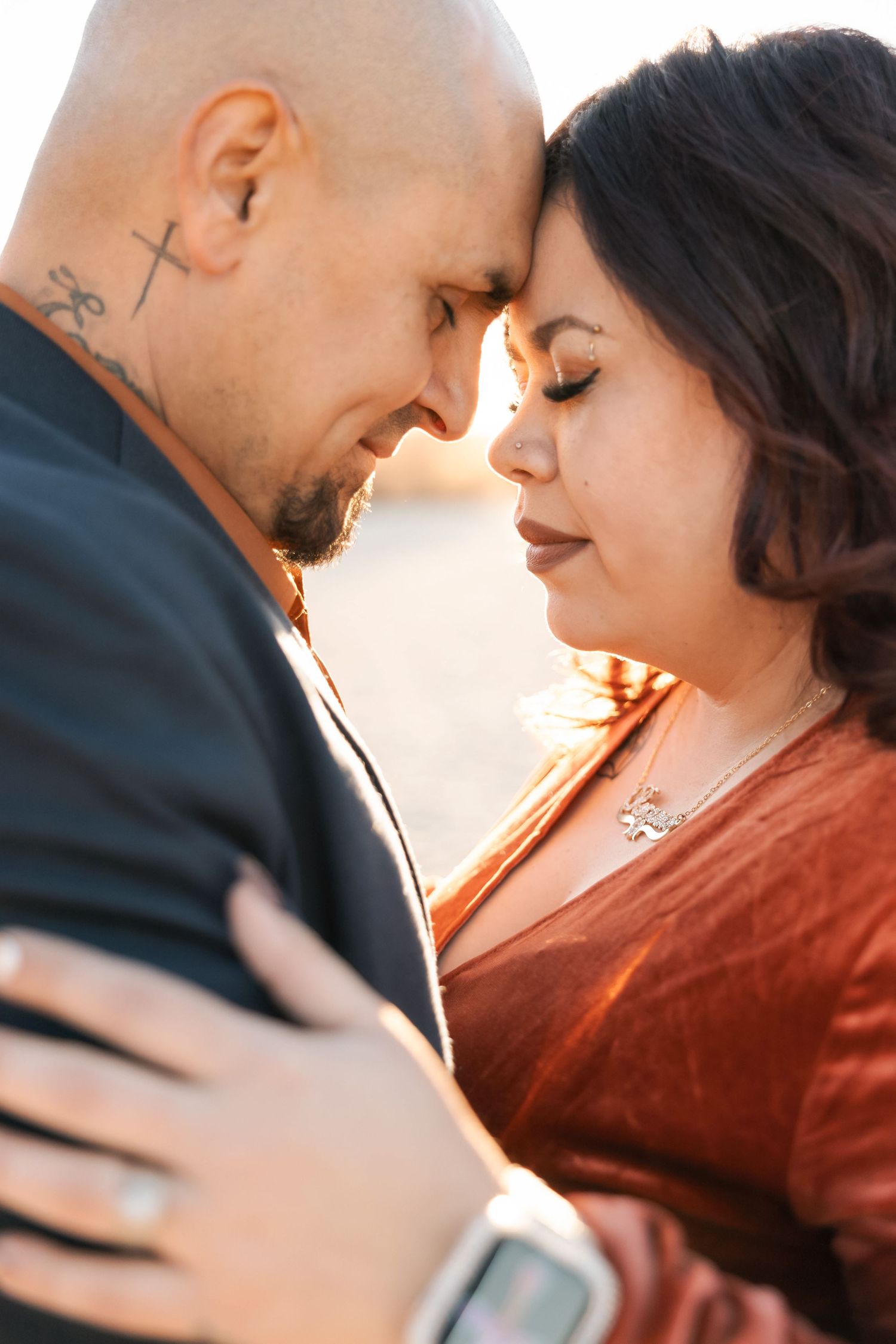A romantic couple in formal attire share an intimate moment against a warm sunset background.