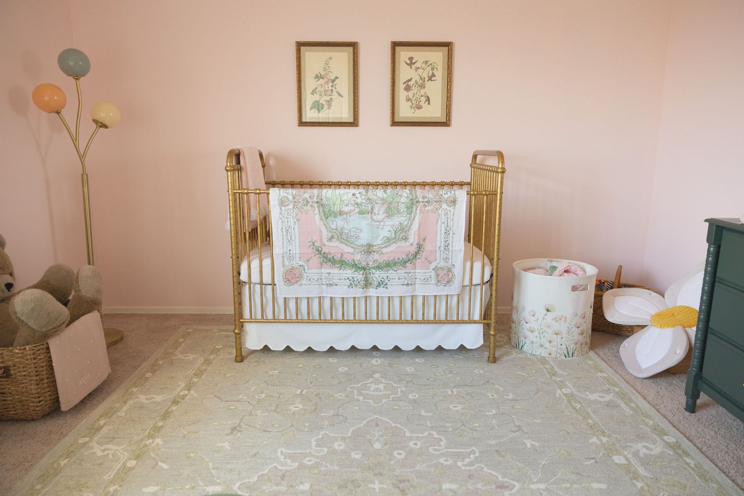 Gold crib with floral bedding on a sage green rug in a pink nursery with botanical art prints on the wall.