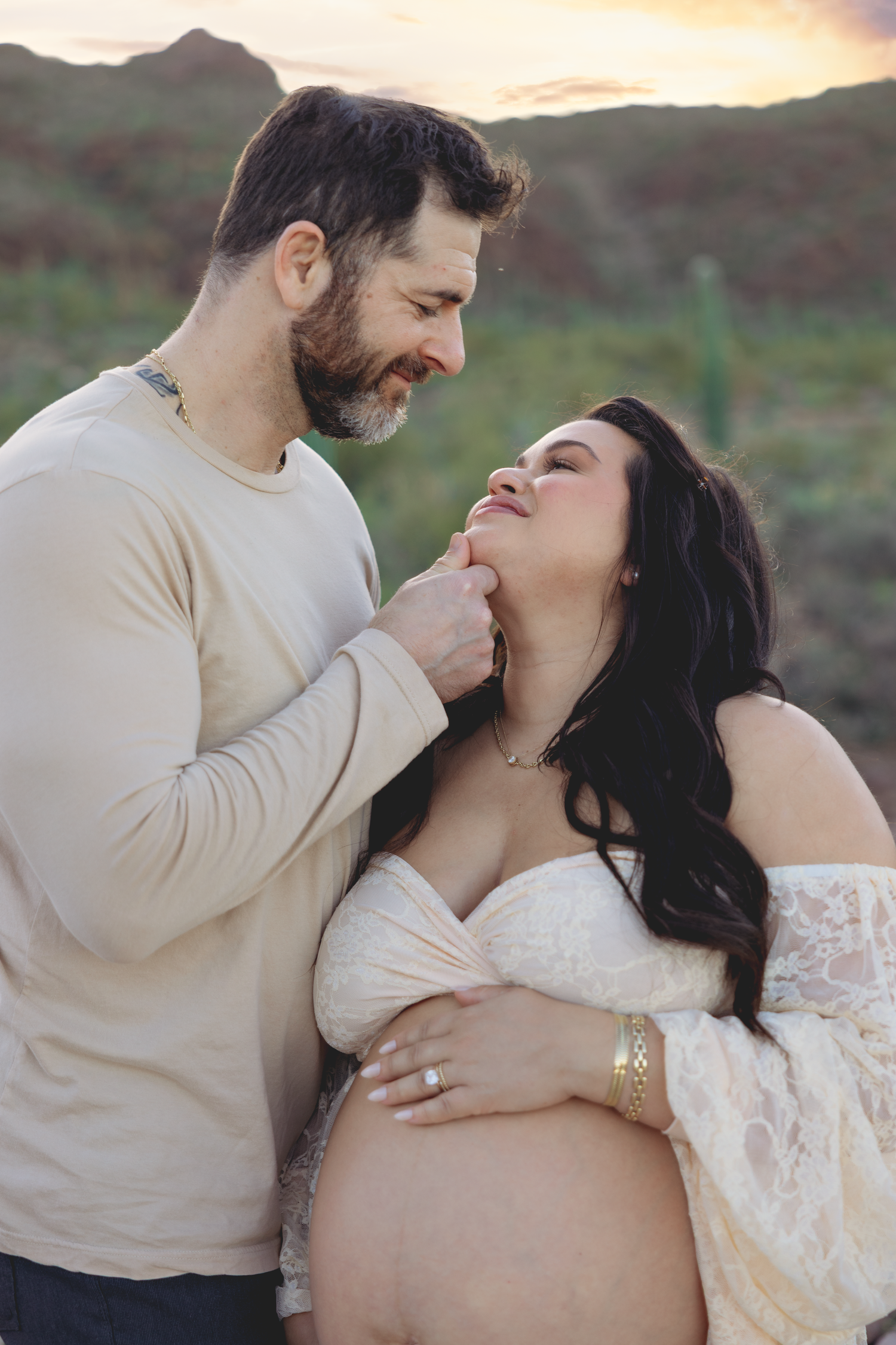 A couple shares a tender moment outdoors at sunset, with the woman's pregnant belly visible in a romantic maternity photo.