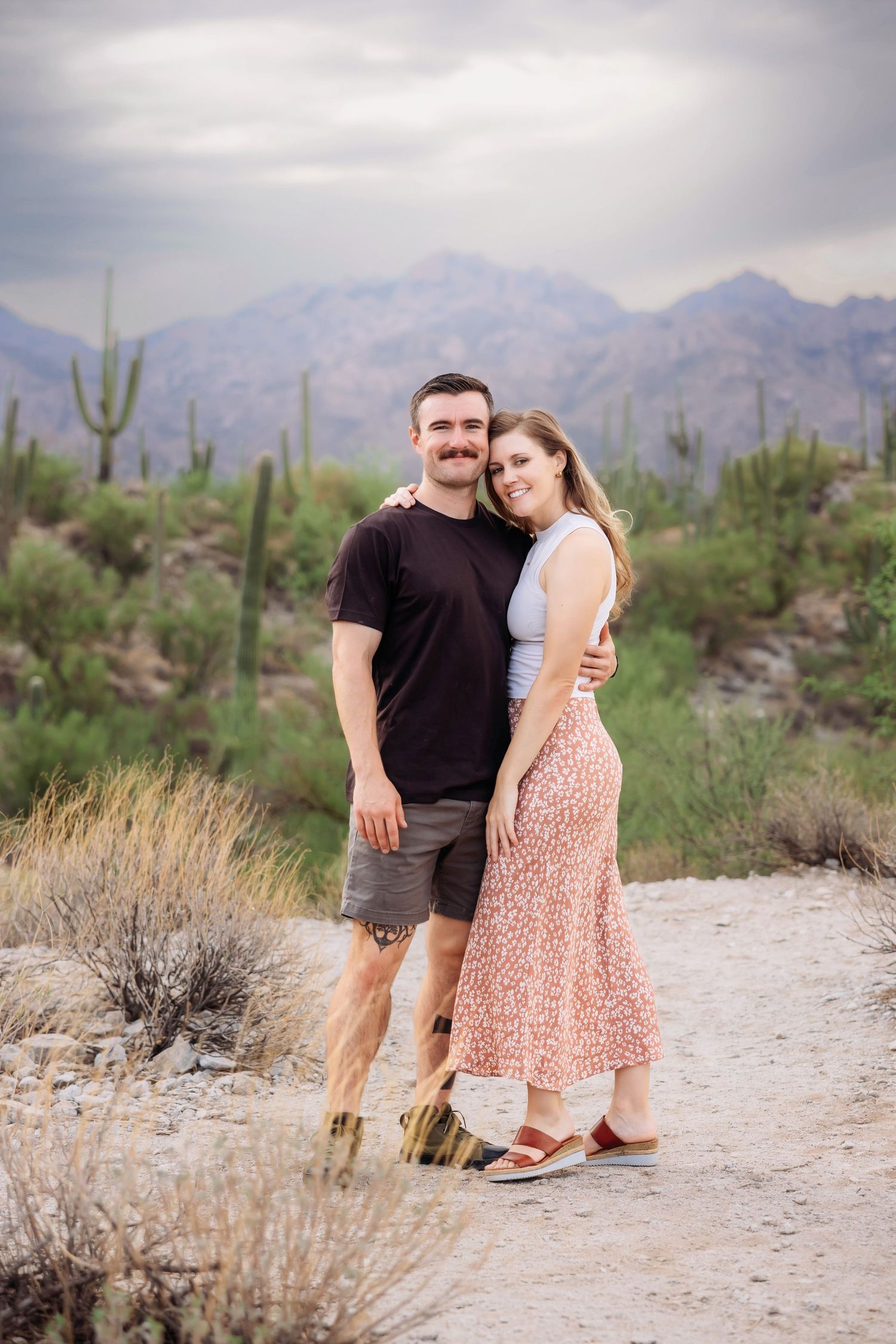 A couple poses together on a desert trail with saguaro cacti and mountains in the background.