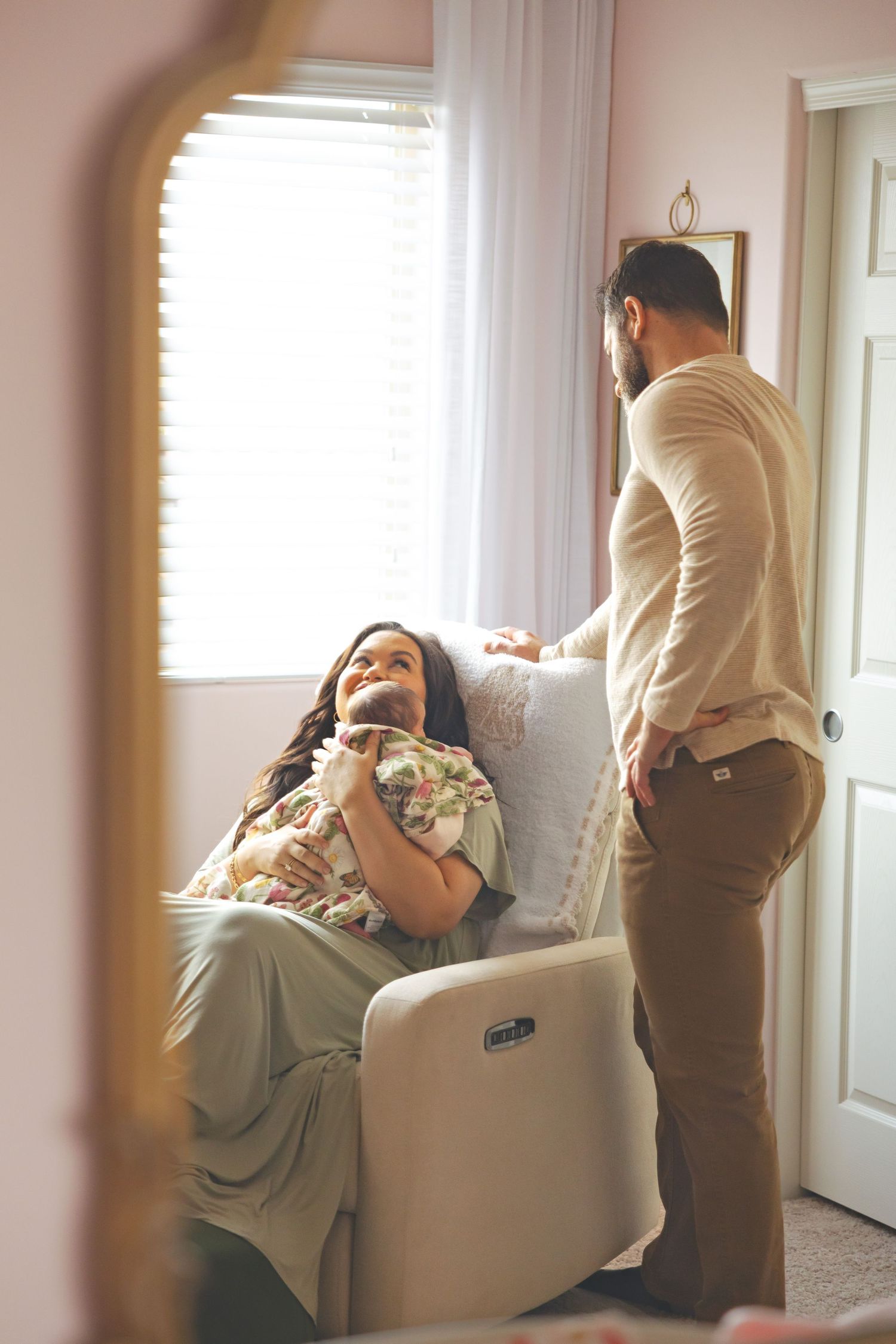 A woman sits in a recliner holding a newborn while a man stands beside her in a softly lit nursery.