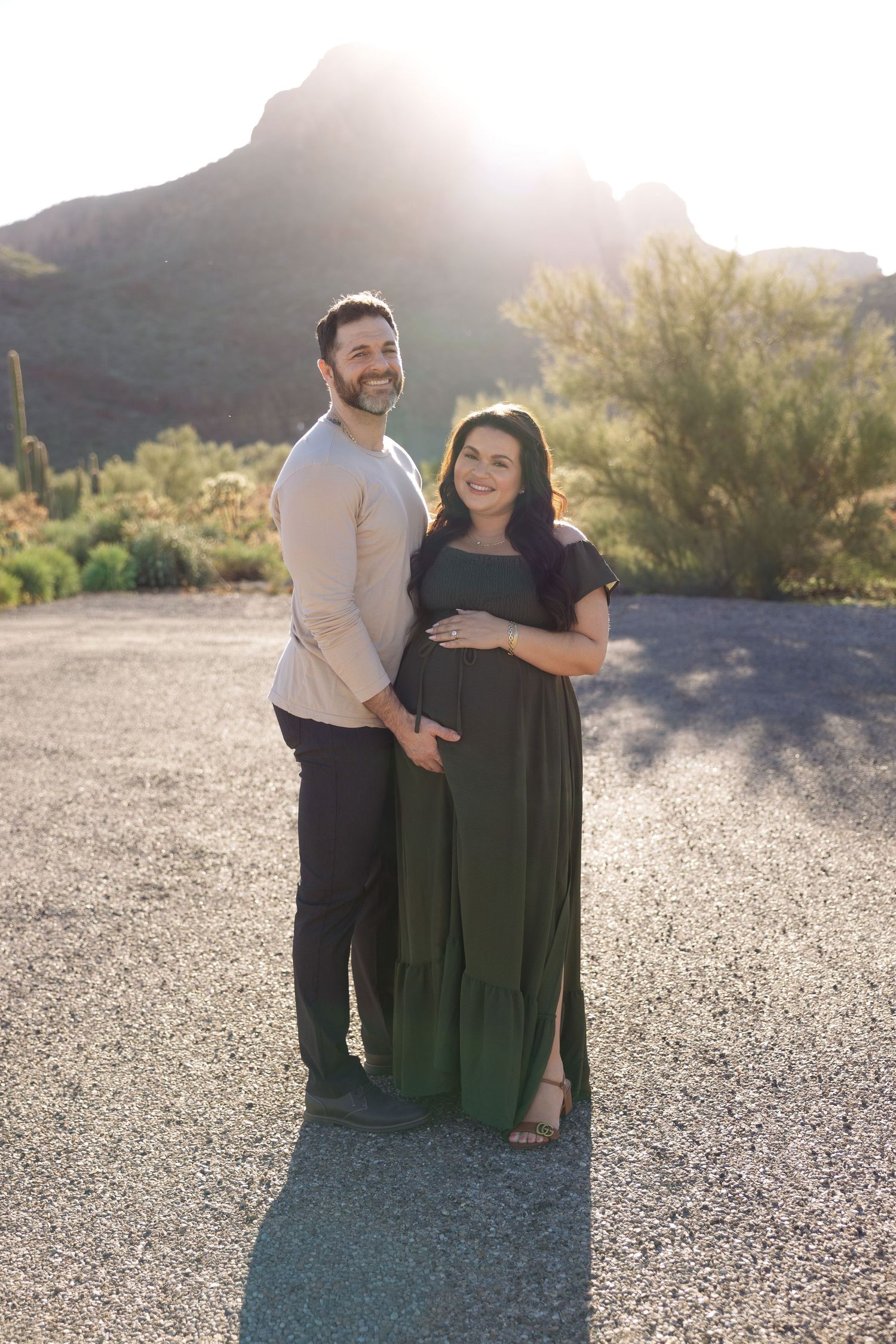 Couple poses outdoors for maternity photos with desert mountain backdrop at golden hour, woman in green maxi dress.