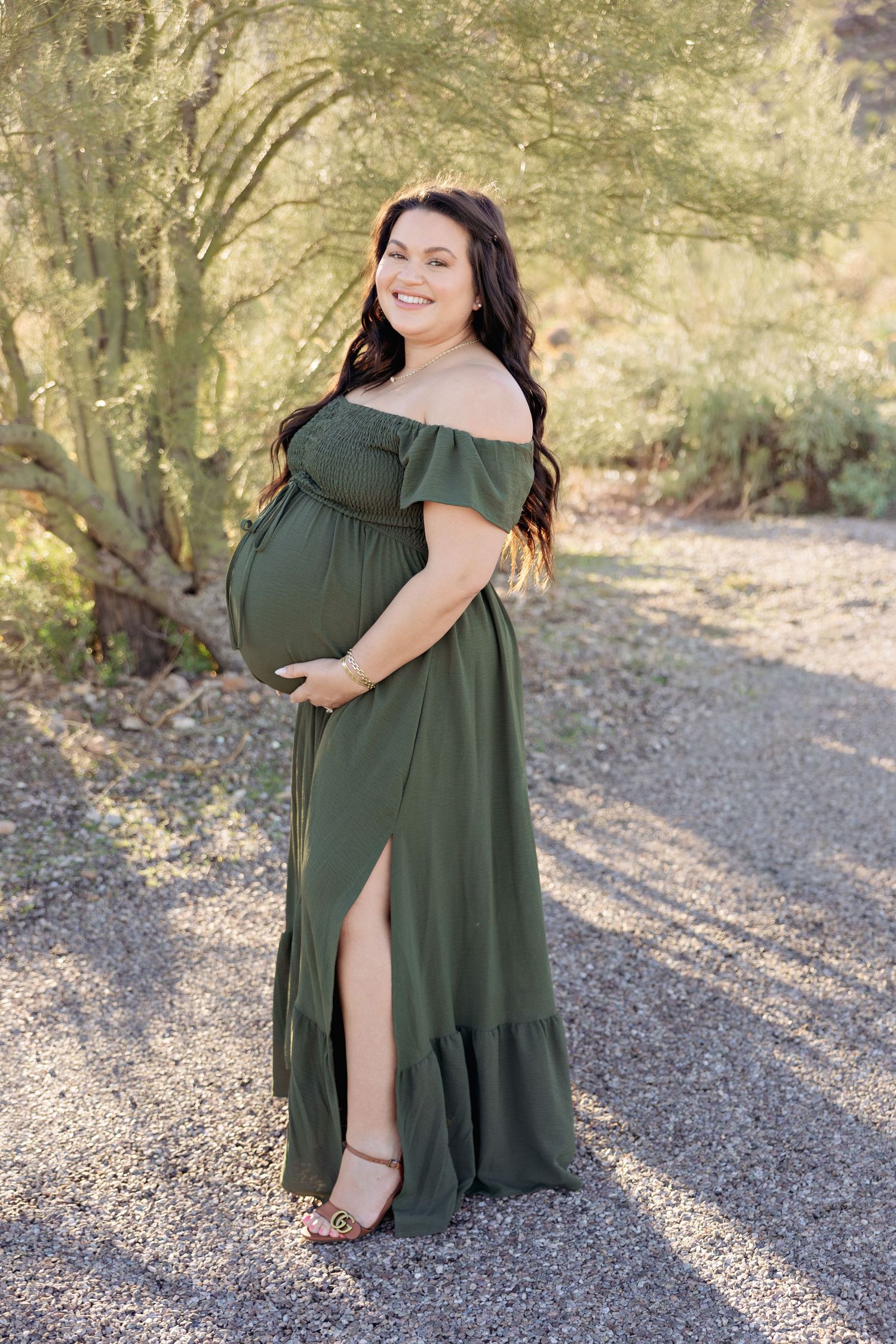 A flowing olive green maxi dress is showcased in an outdoor setting with dappled sunlight and natural surroundings.