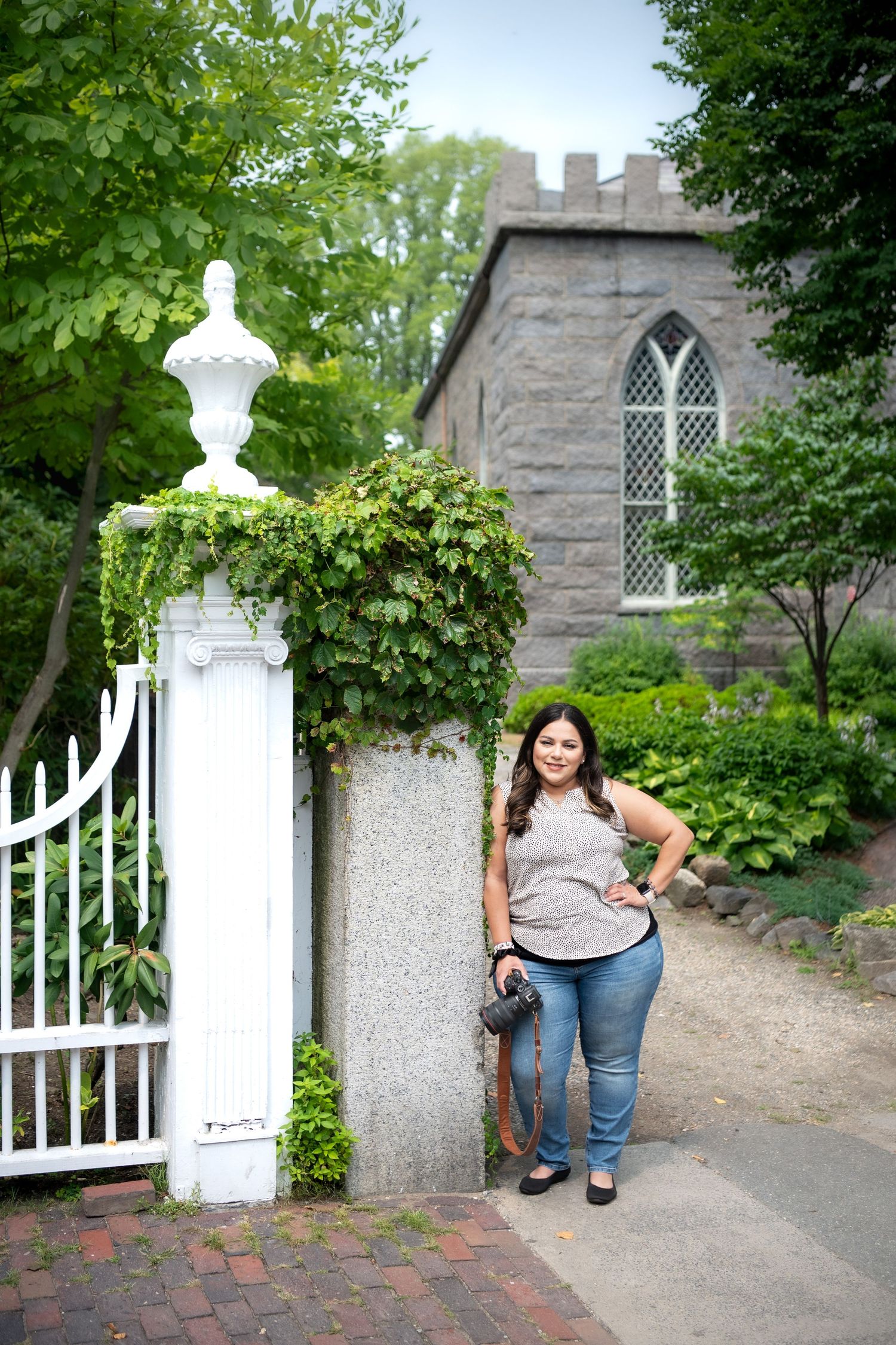 A lifestyle portrait by an ornate stone gate post with ivy near a gothic stone church building.