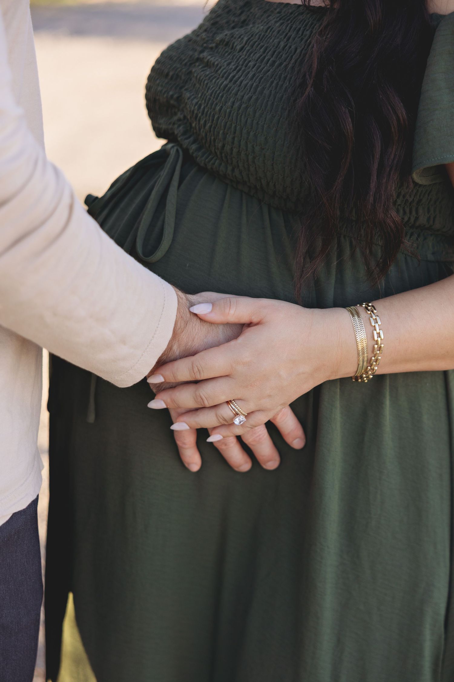 Couple holding hands over pregnant belly, woman wearing diamond ring and gold bracelet in green maternity dress.