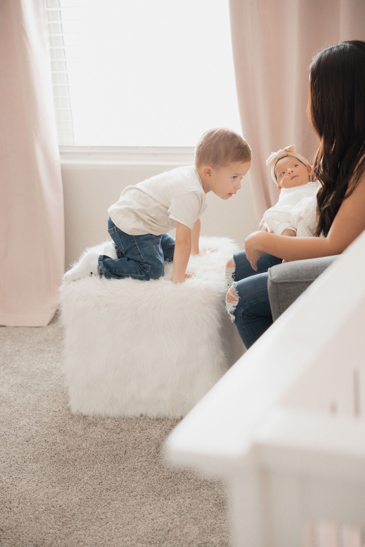 A toddler boy leans over a fluffy white ottoman toward a mother holding her newborn baby in a soft pink nursery.