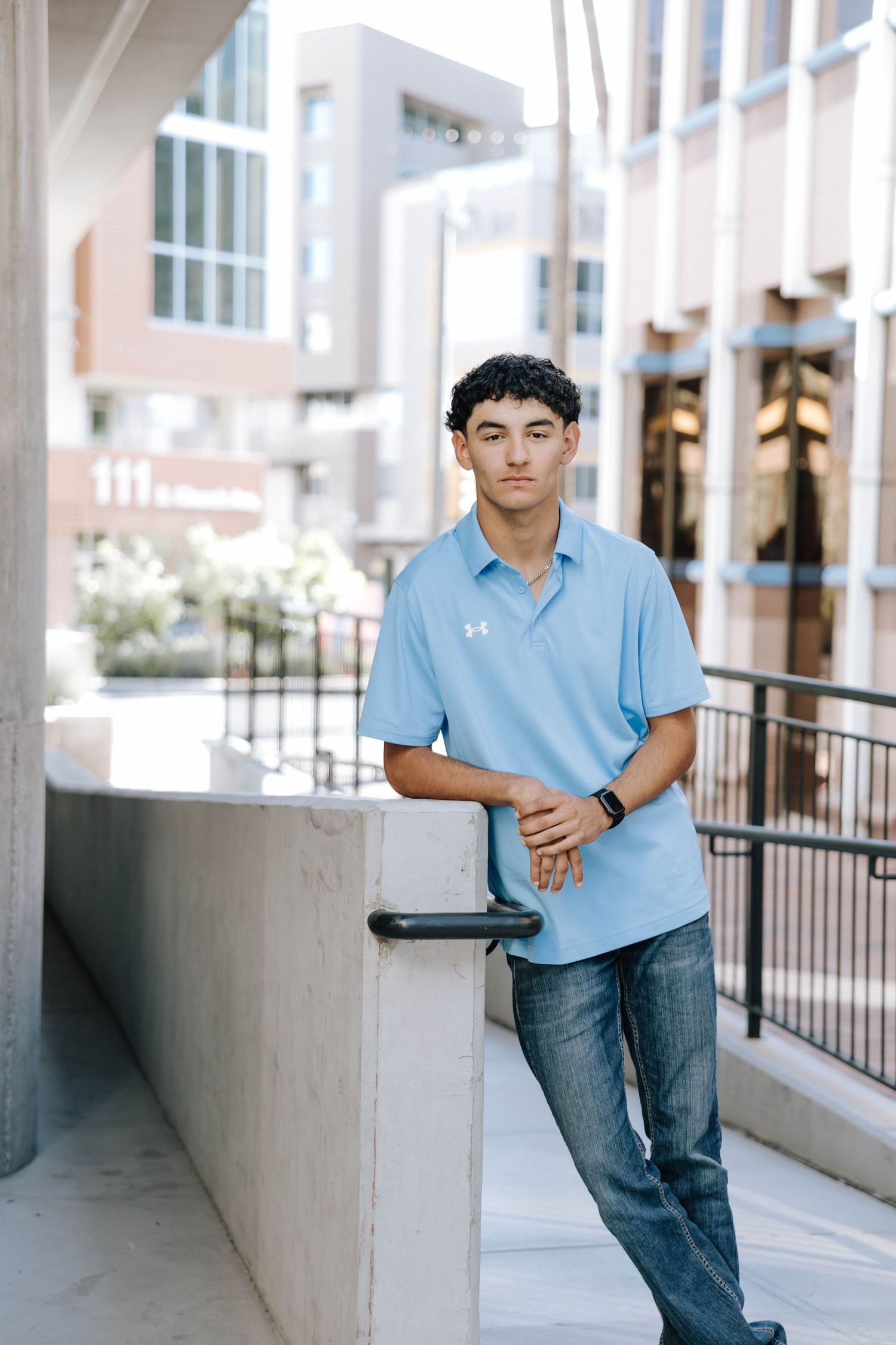 Student in light blue polo shirt and jeans poses casually against wall in outdoor campus setting.