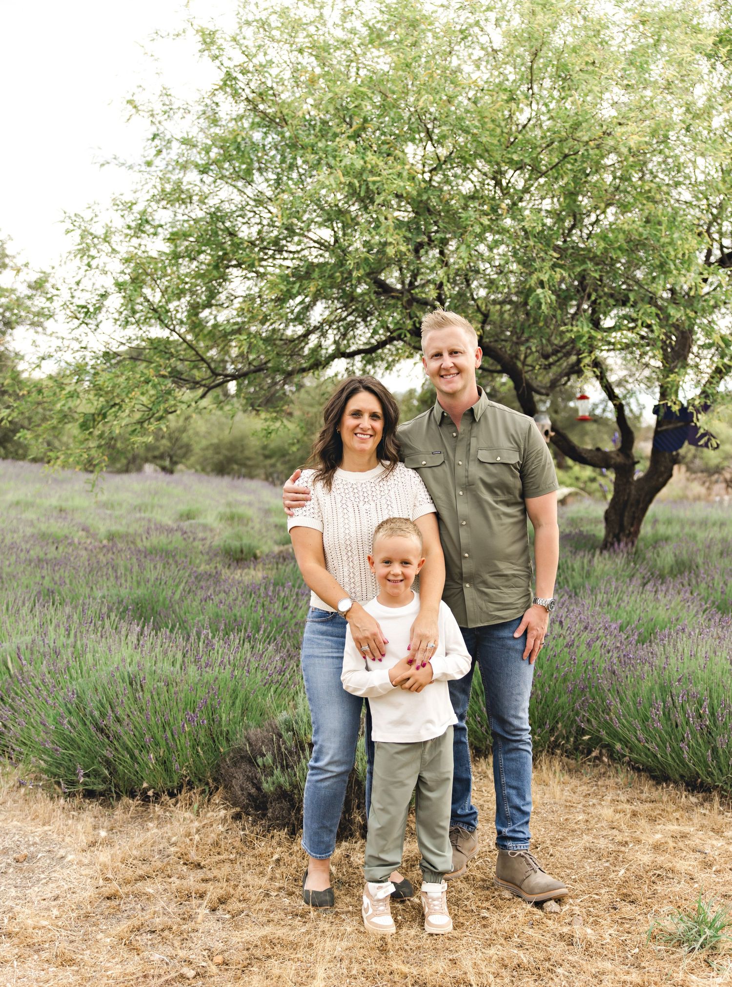 Family of three smiling in a lavender field with a lush green tree backdrop on a cloudy day.