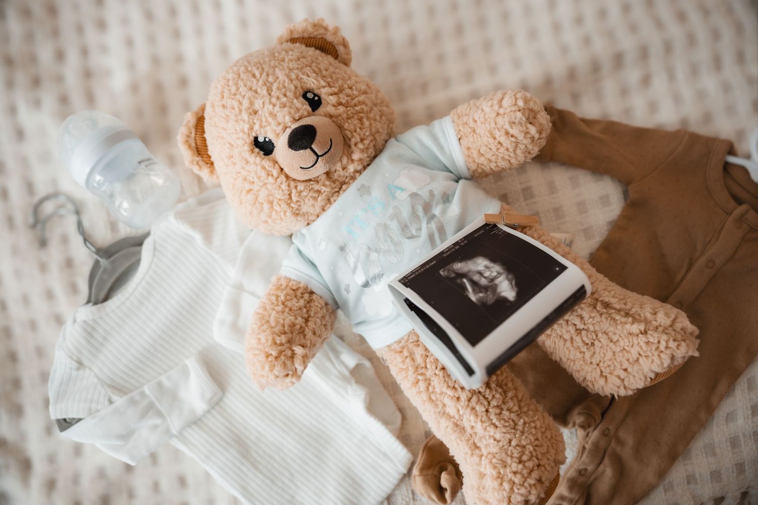 A teddy bear lies next to baby clothes and an ultrasound photo on a white textured surface.