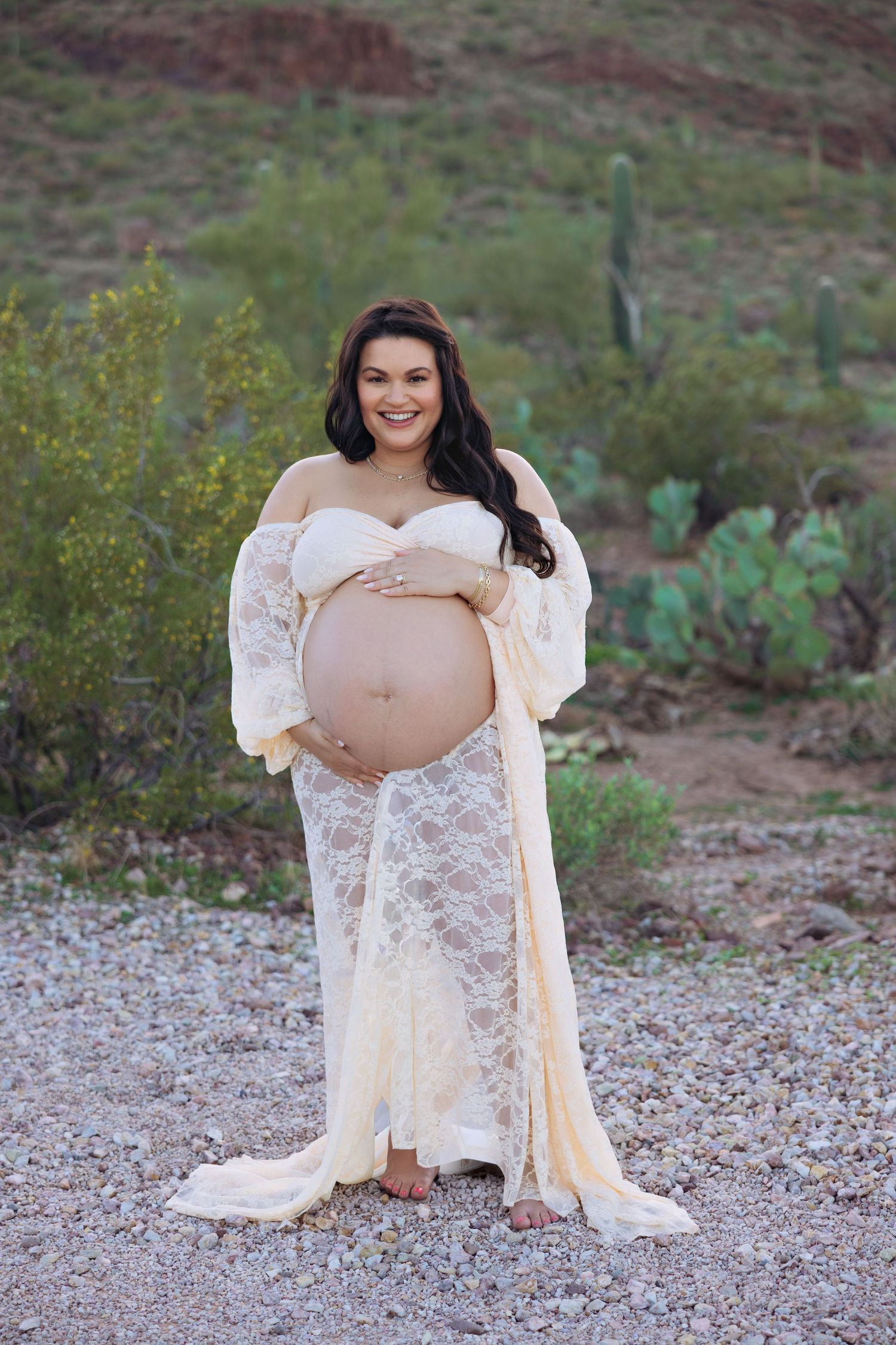 Smiling pregnant woman in lace maternity gown cradling baby bump outdoors in desert landscape with cacti.